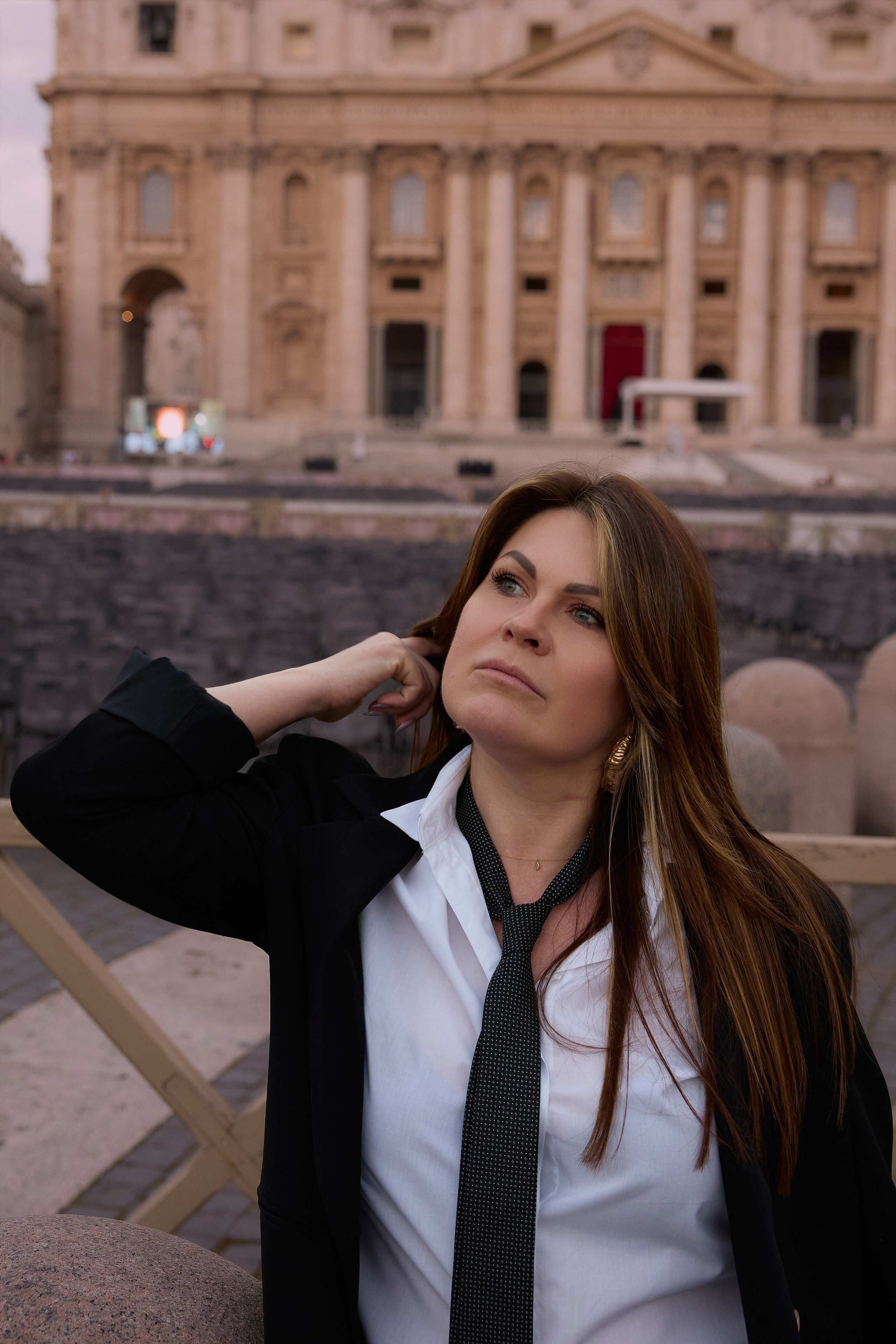 Портрет женщины в черном пиджаке, белой рубашке и свободном темном галстуке, стоящей на открытом воздухе на фоне классического фасада собора Святого Петра или аналогичной достопримечательности.