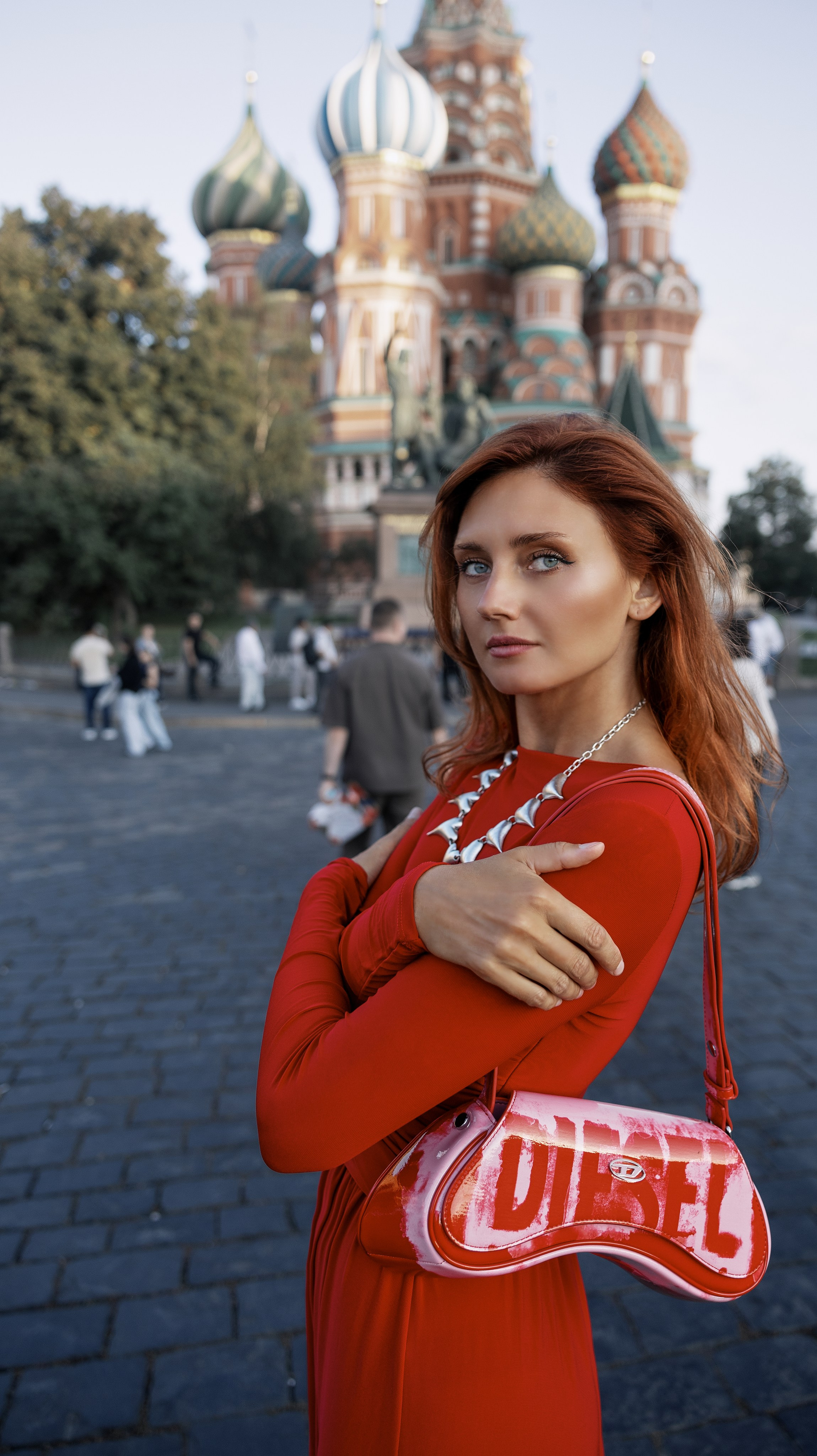 «Модное фото женщины в красном платье с длинными рукавами и красной лакированной сумке Diesel через плечо с бело-розовым логотипом в стиле граффити. Она стоит на мощёной площади перед собором Василия Блаженного в Москве с его разноцветными куполами, видными на размытом фоне».