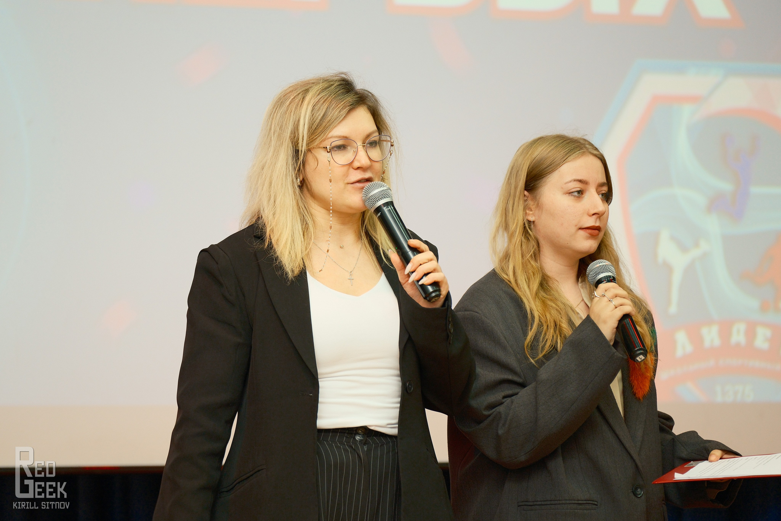 Две ведущие-блондинки на сцене: одна объявляет программу, вторая держит лист выступлений.