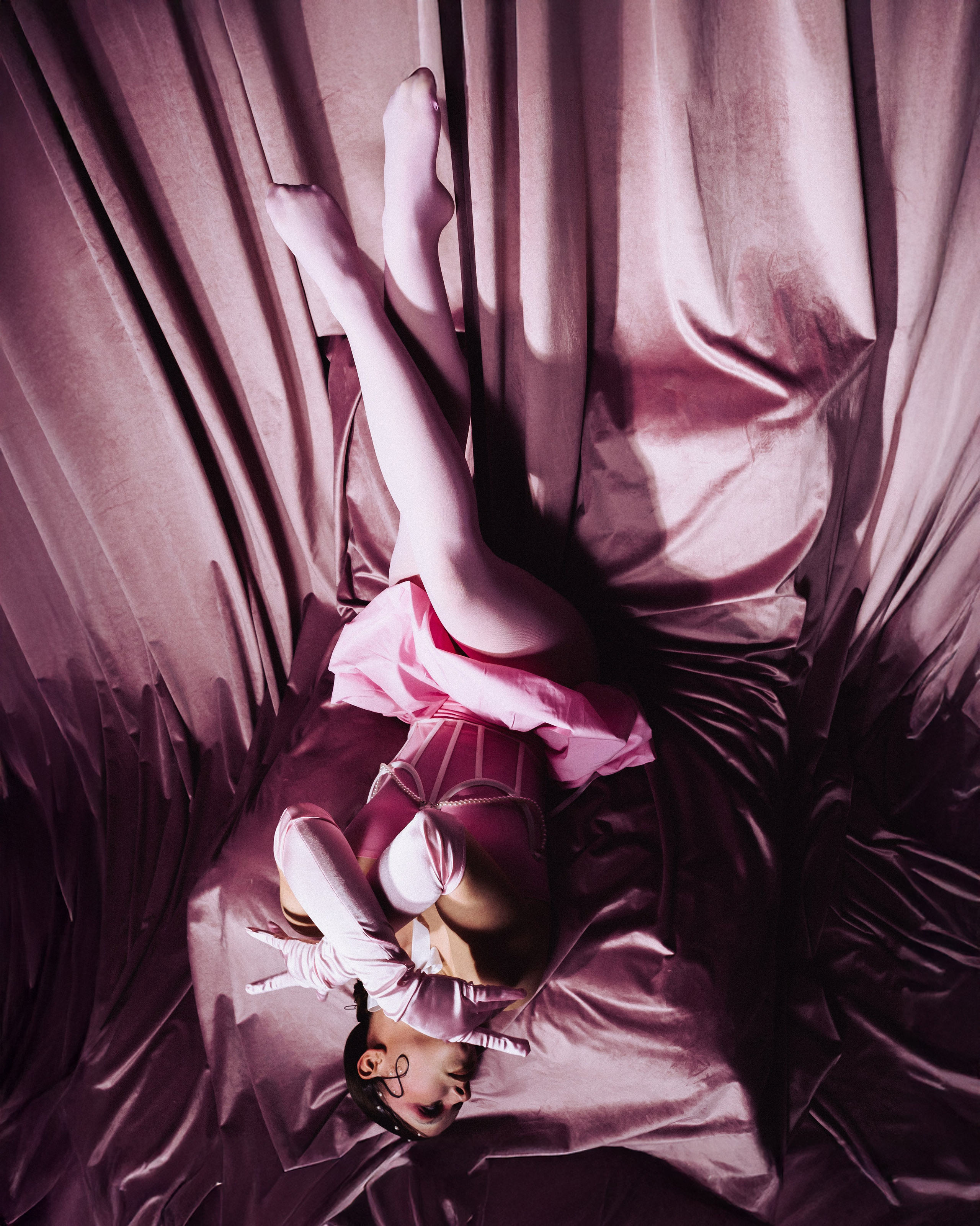 Фотограф Александра Овечкина Пенза: вертикальный ракурс, модель лежит на спине с поднятыми вверх ногами, утопая в складках розового бархата.