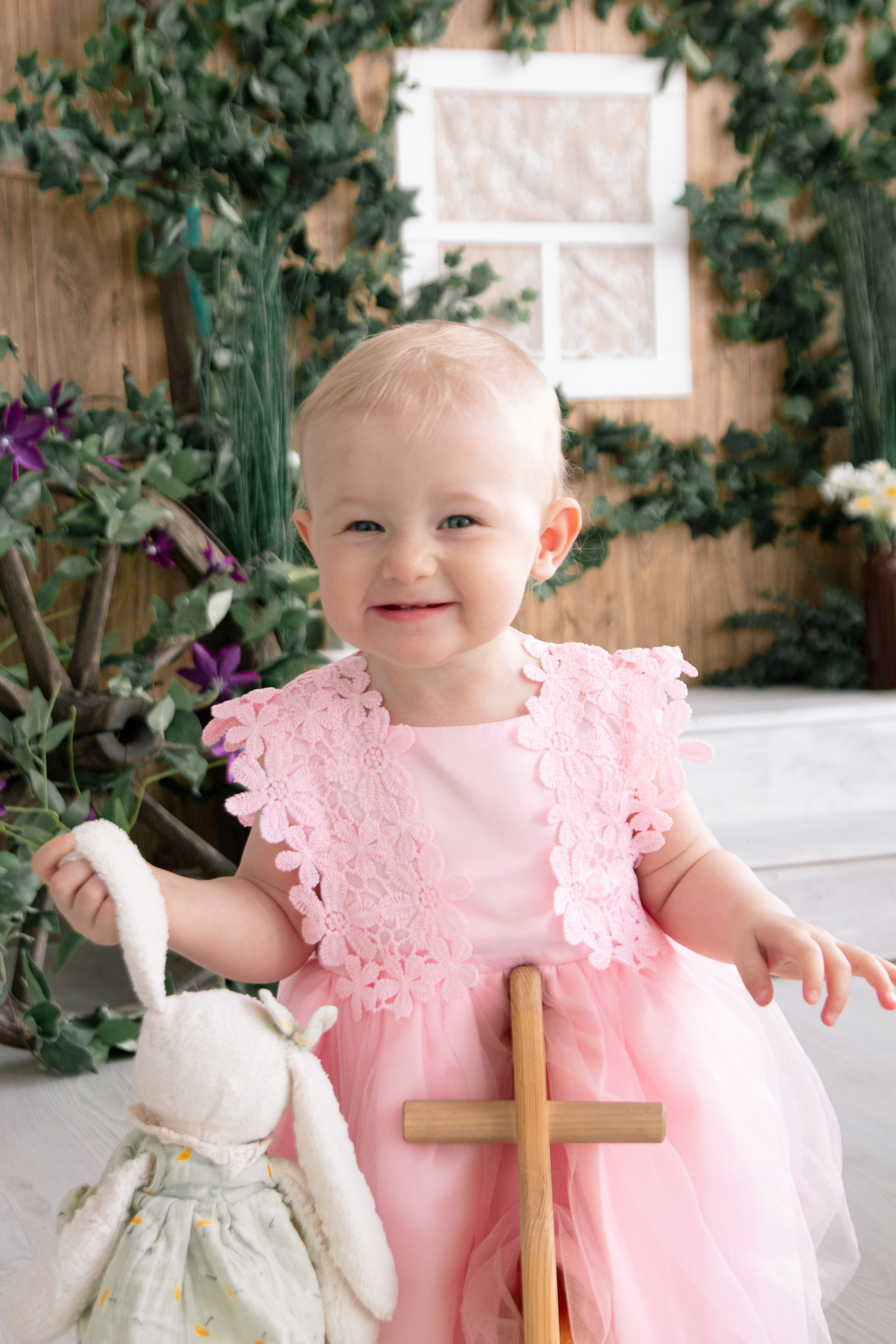 Малышка в розовом платье с игрушкой в руках на фоне зеленых листьев