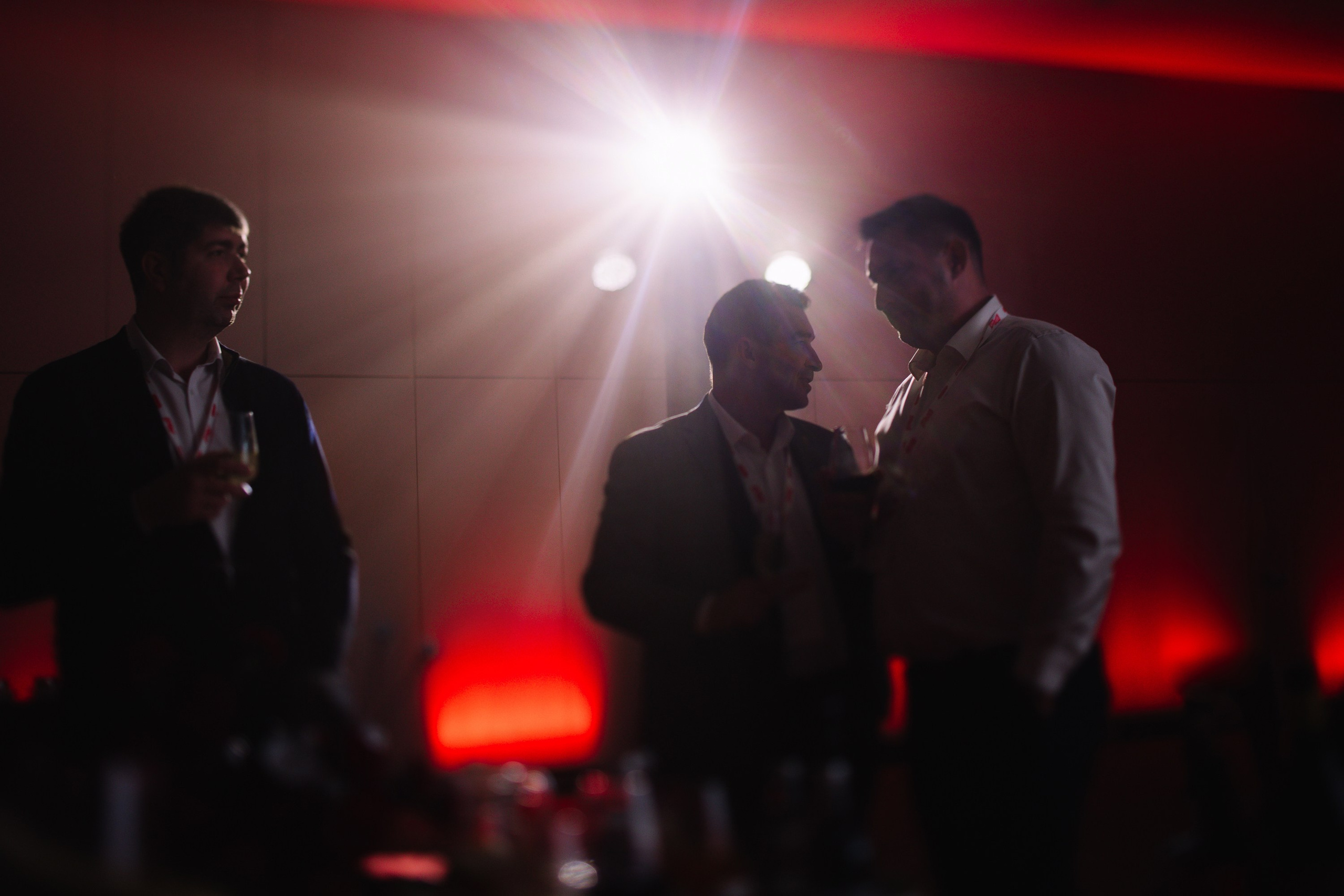 Общение: Трое мужчин в деловых костюмах ведут беседу в лаунж-зоне мероприятия в мягком вечернем свете.