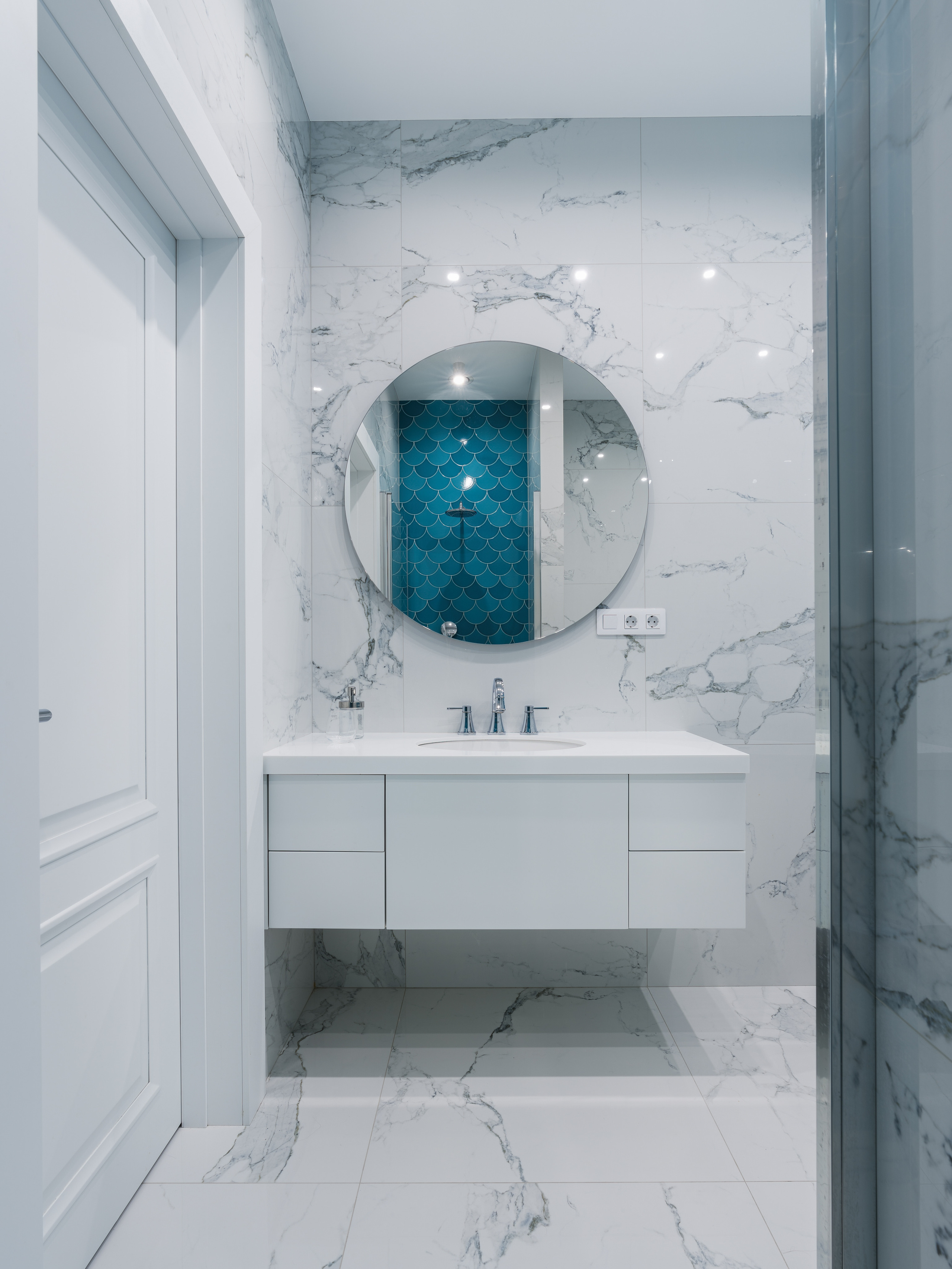 Современная ванная с крупным круглым зеркалом, мраморной плиткой и белой подвесной тумбой под раковину, акцентная стена с синей плиткой чешуя; интерьер от Евгении Княжевой, интерьерный фотограф Сергей Денисенко.
