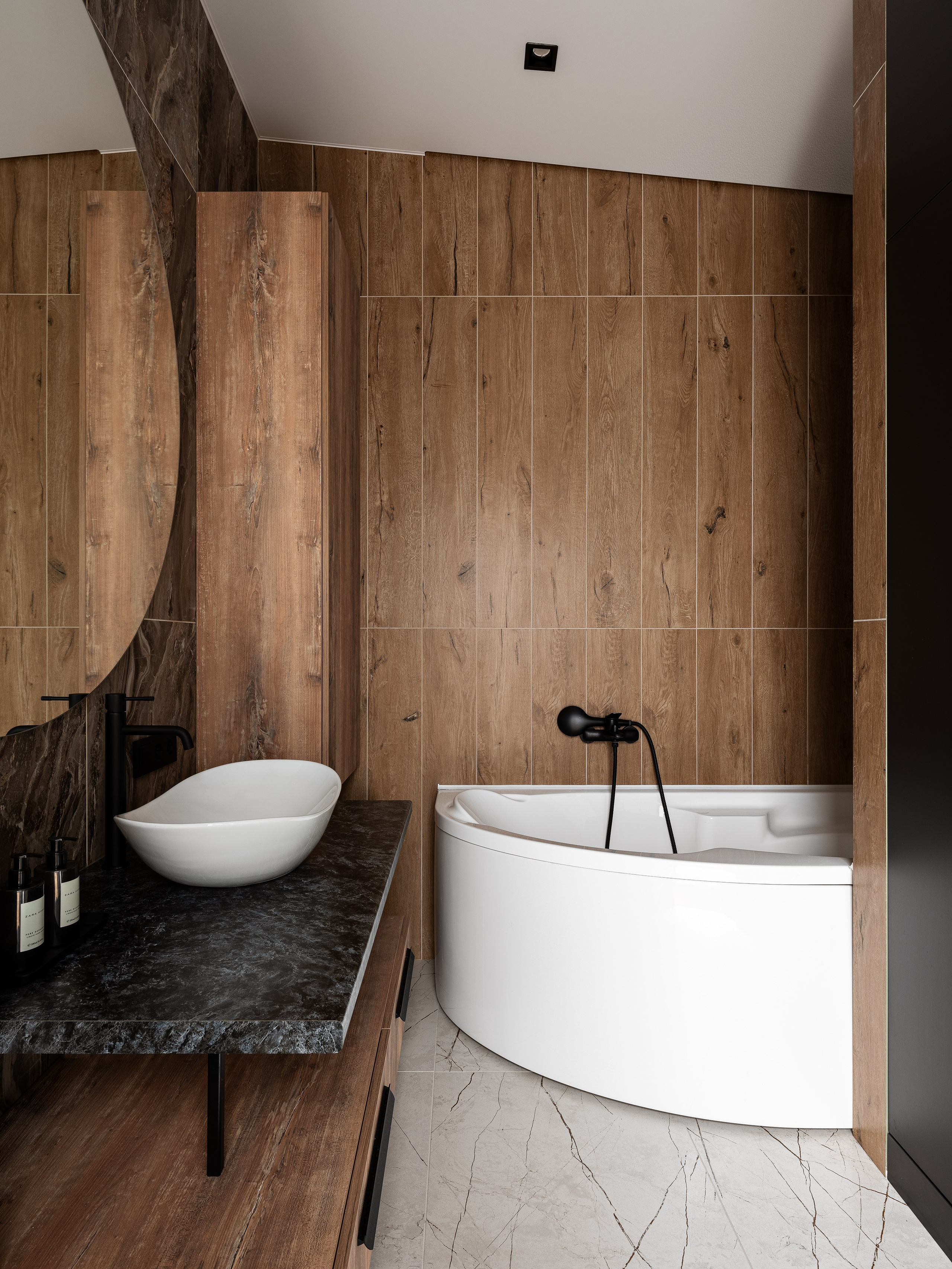 Ванная комната с угловой белой ванной, деревянной отделкой стен, черной столешницей с дизайнерской раковиной и встроенной подсветкой; современный проект Екатерины Мищенко, фотограф Сергей Денисенко.