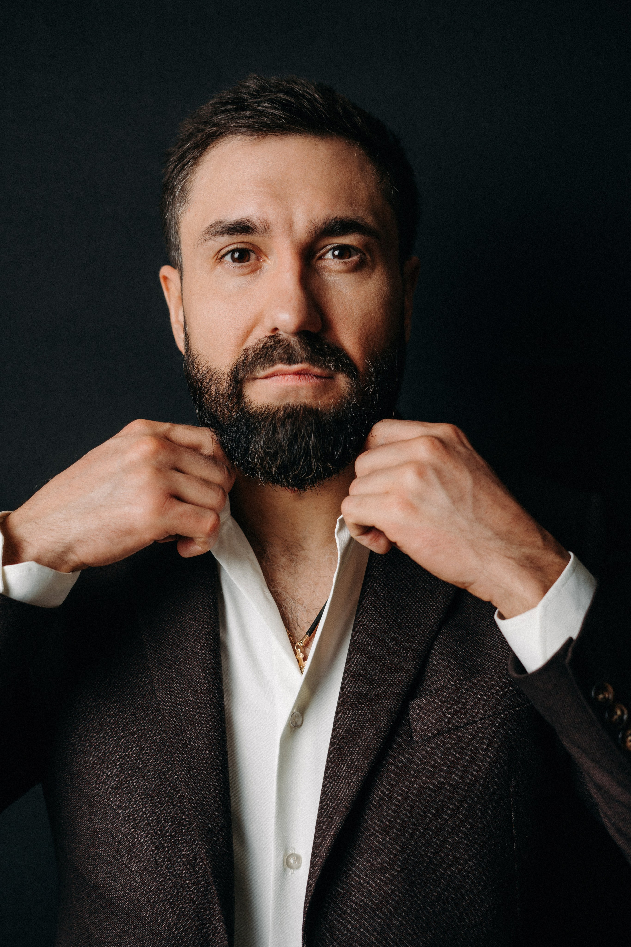 Портрет мужчины брюнета с бородой в пиджаке на черном фоне поправляет ворот рубашки