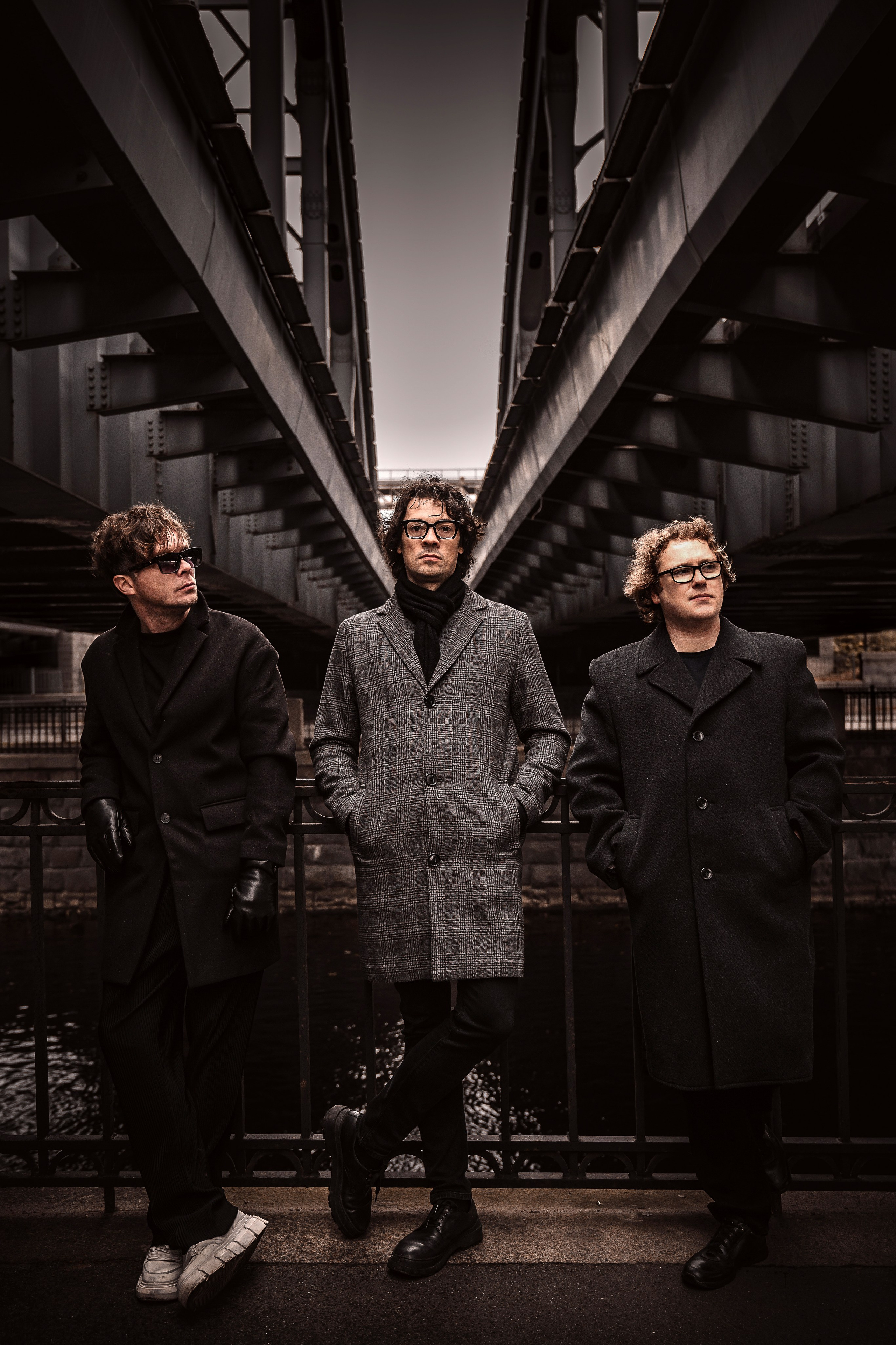Радио Сатурн. Три участника российской рок-группы в тёмных пальто, очках и кедах стоят на  мосту над рекой в промышленной зоне, атмосферный портрет в серых тонах»