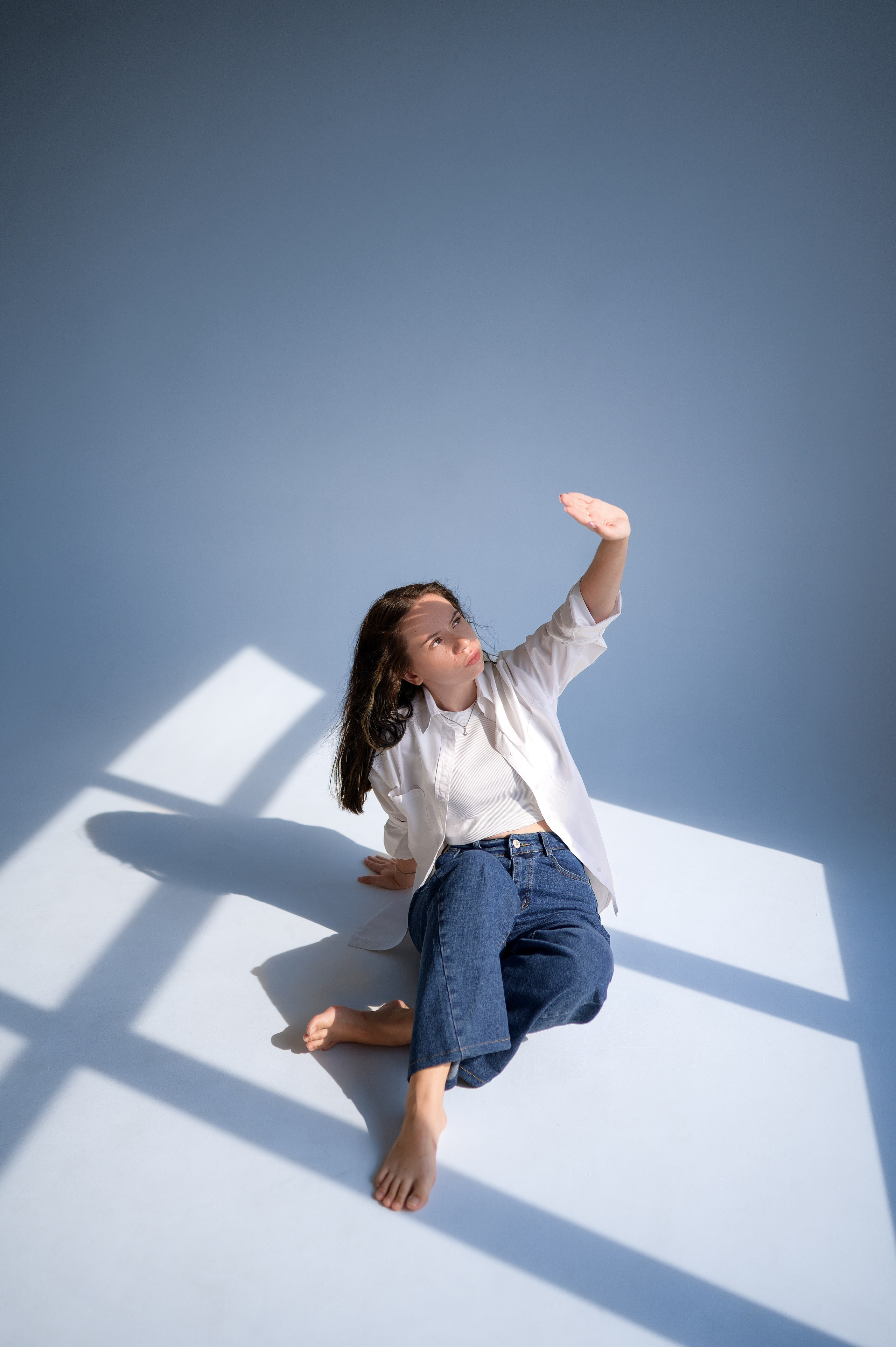 Минималистичная съёмка в студии: девушка в повседневной одежде (рубашка, джинсы) на белой циклораме. Лучи света создают геометричные тени. Волгодонск