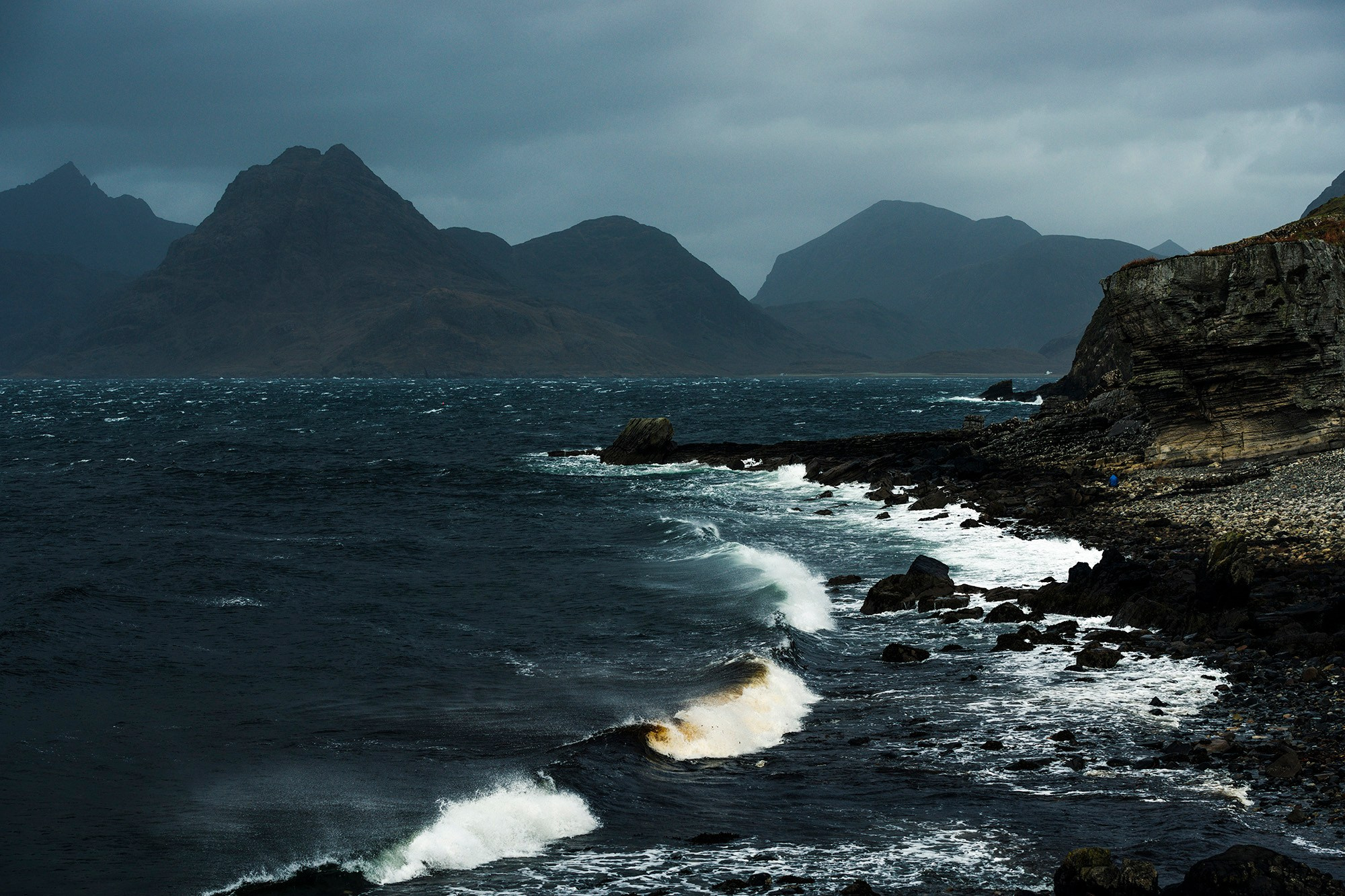 Суровый прибрежный пейзаж острова Скай, Шотландия, с бурным темным морем, разбивающимися волнами и большими горами на заднем плане под пасмурным небом.