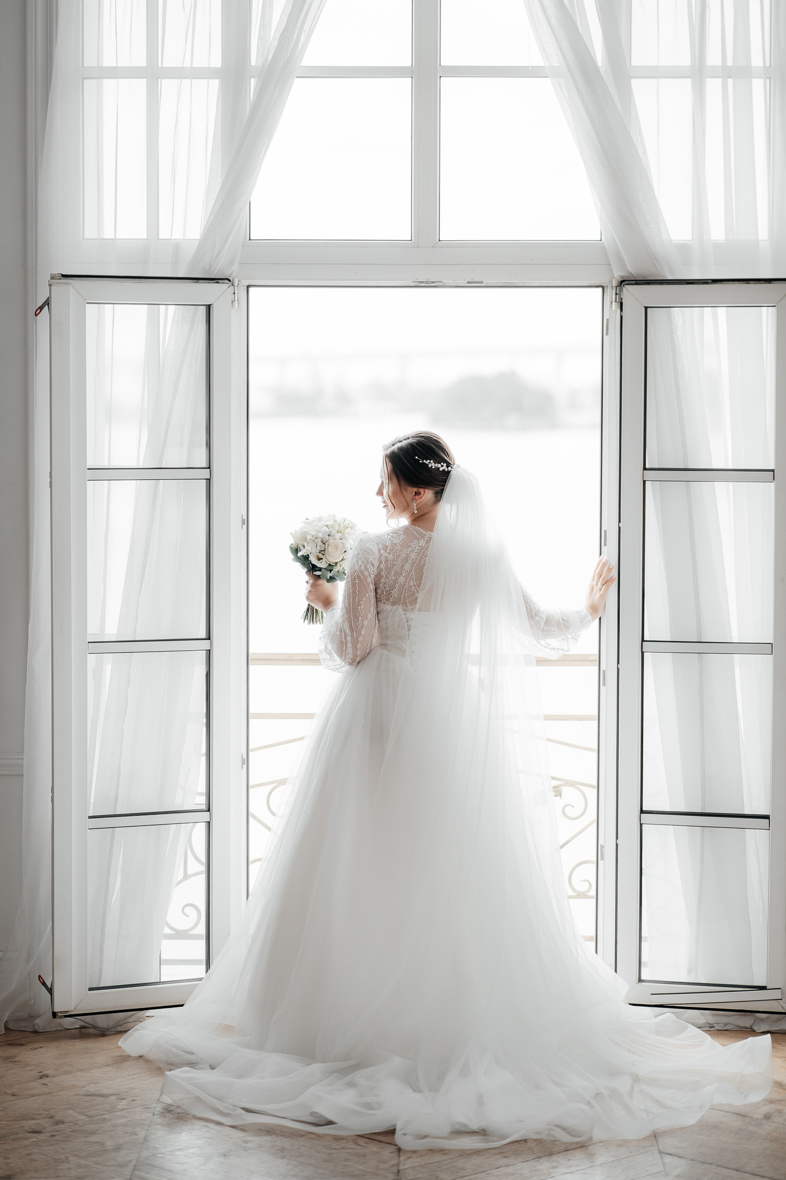 Невеста в белом платье стоит около окна во весь рост