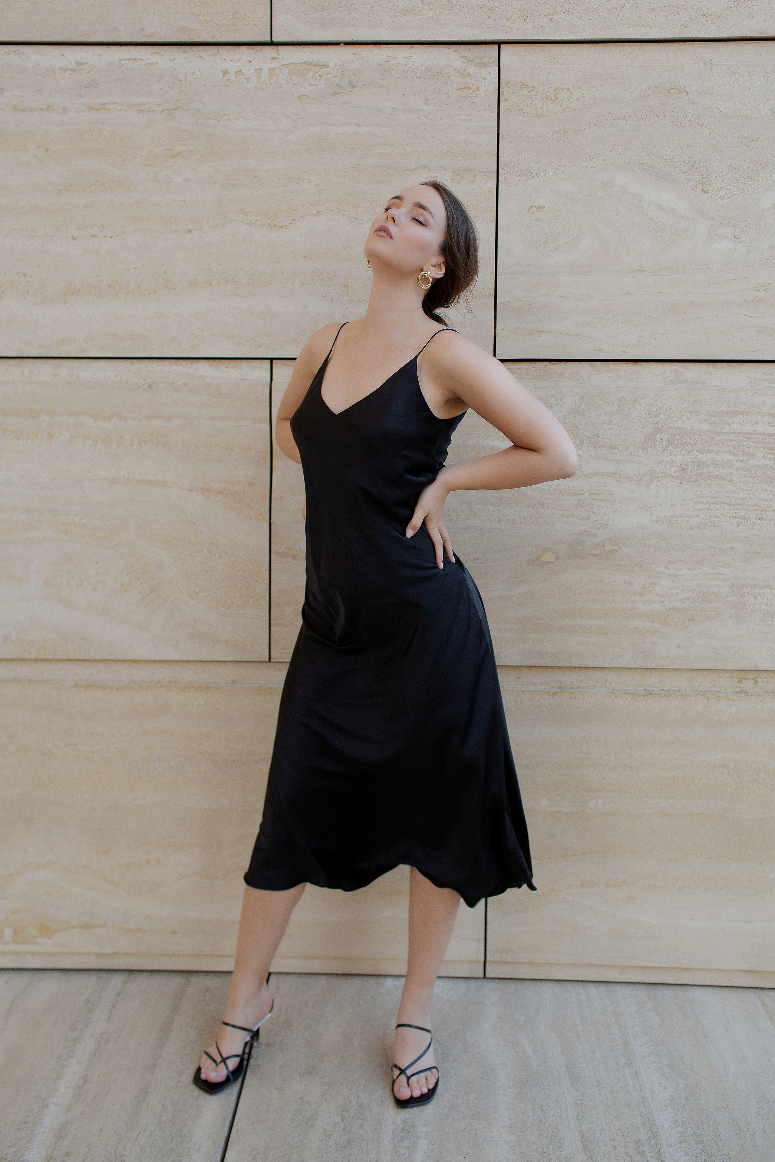 ростовое фото девушки в черном атласном платье на фоне бежевой каменной стены