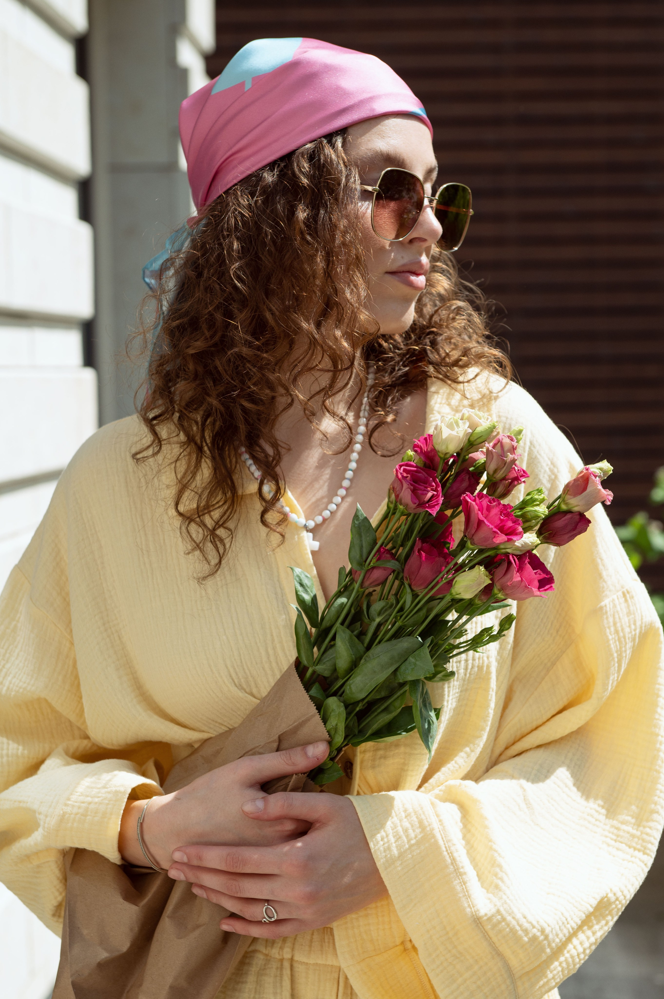 Красивая девушка в дизайнерской одежде и шелковом дизайнерском платке стоит с букетом цветов на фоне стены