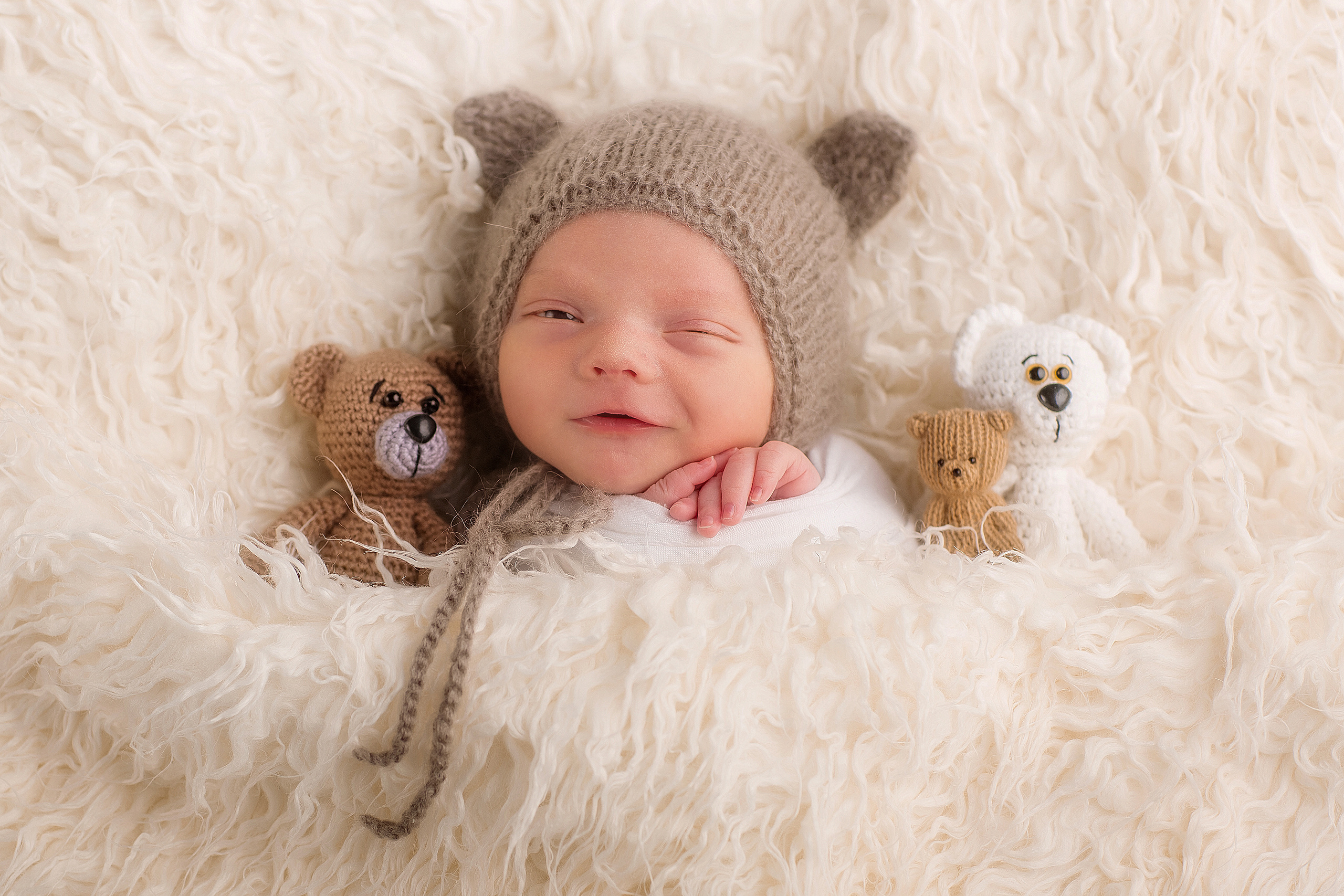 портрет новорожденного малыша  в серой шапке с ушками ручки под подбородком улыбается и подмигивает