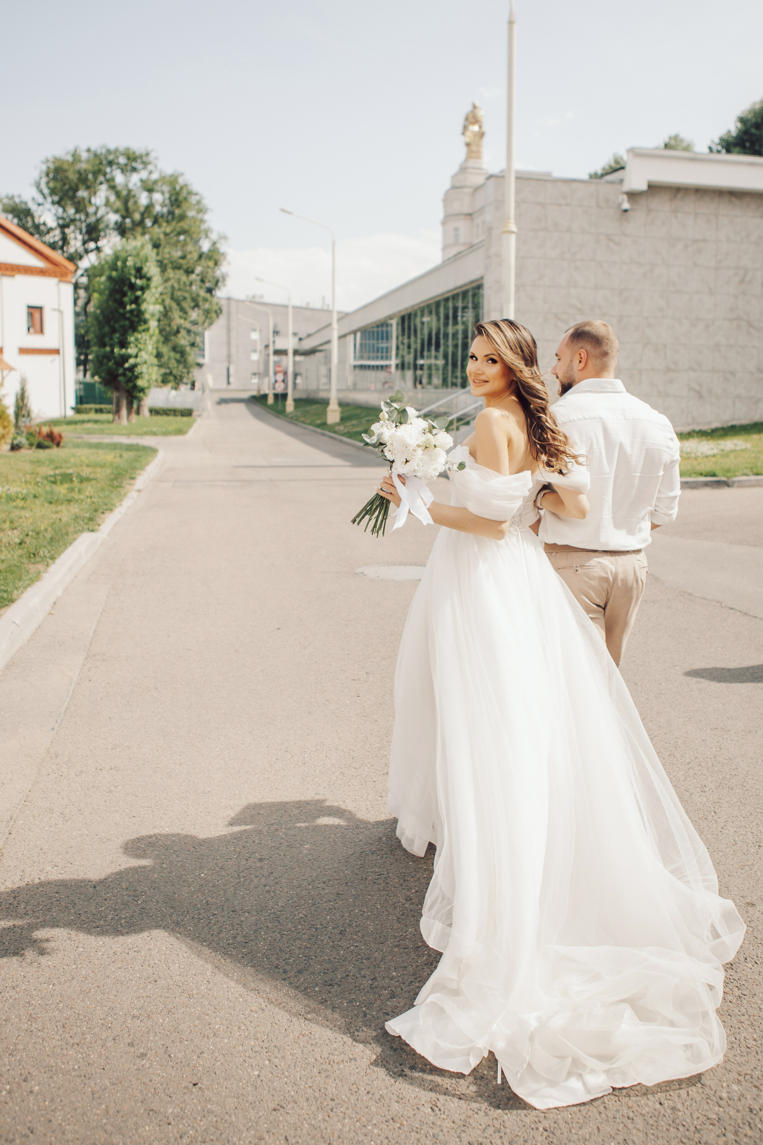 Невеста в длинном белом платье идет по ВДНХ в солнечную погоду
