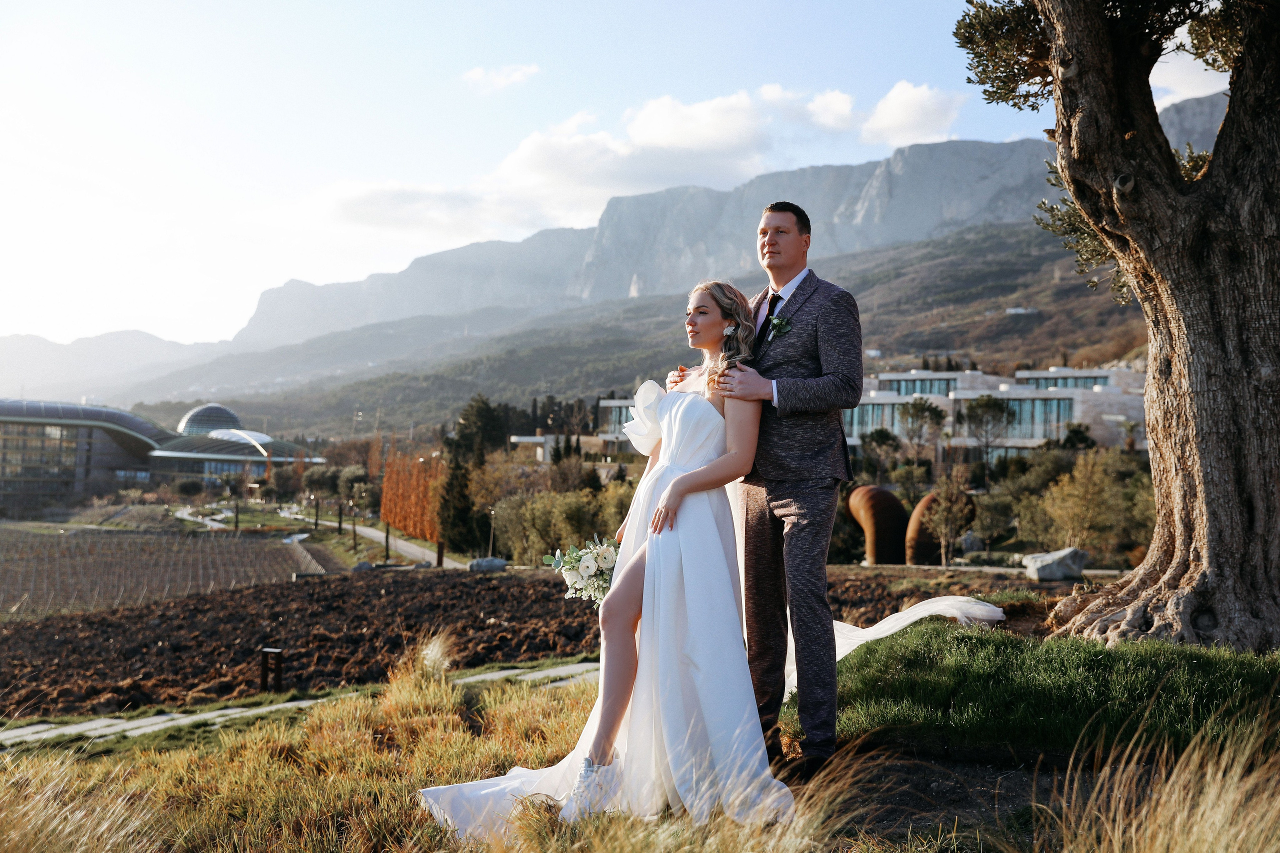 пара в свадебных нарядах у дерева на фоне отеля Мрия и гор в Крыму