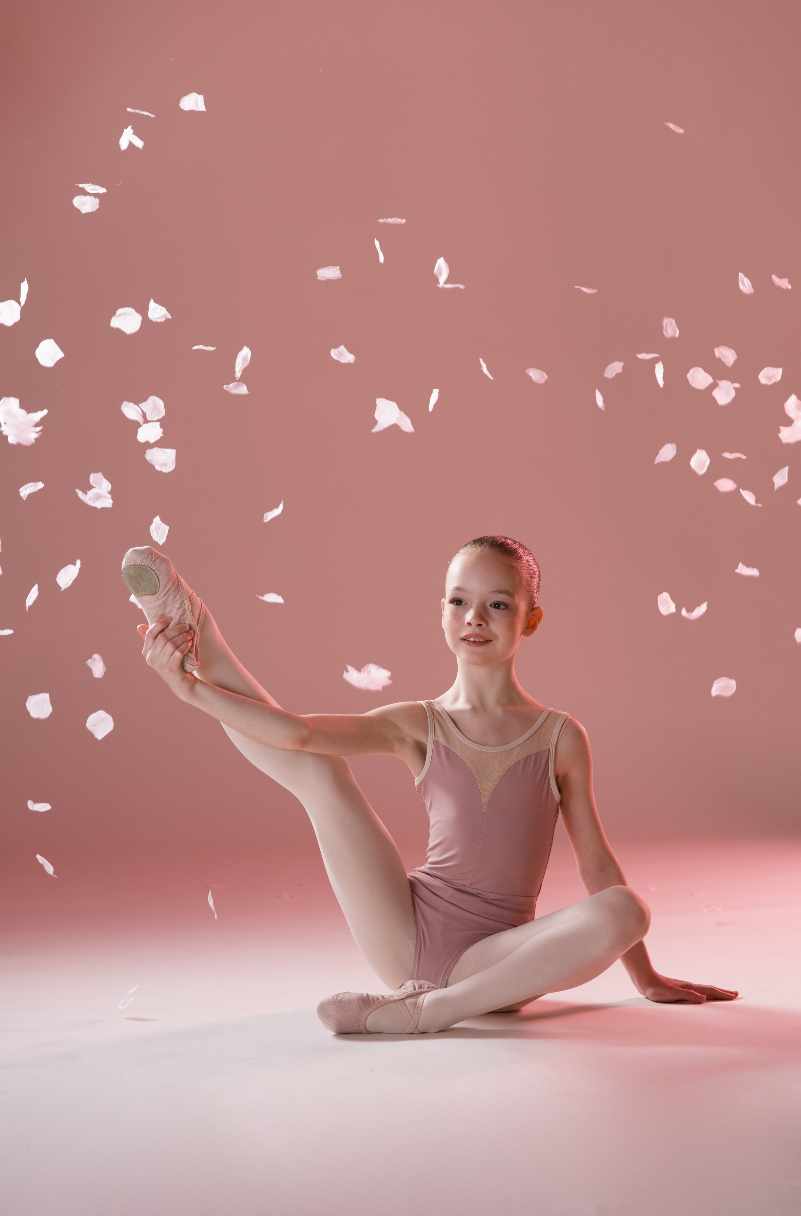 Юная балерина в розовом платье демонстрирует вертикальный шпагат сидя в облаке летящих лепестков. Художественная съемка в стиле спорт-арт с мягким светом, фотограф в Тюмени