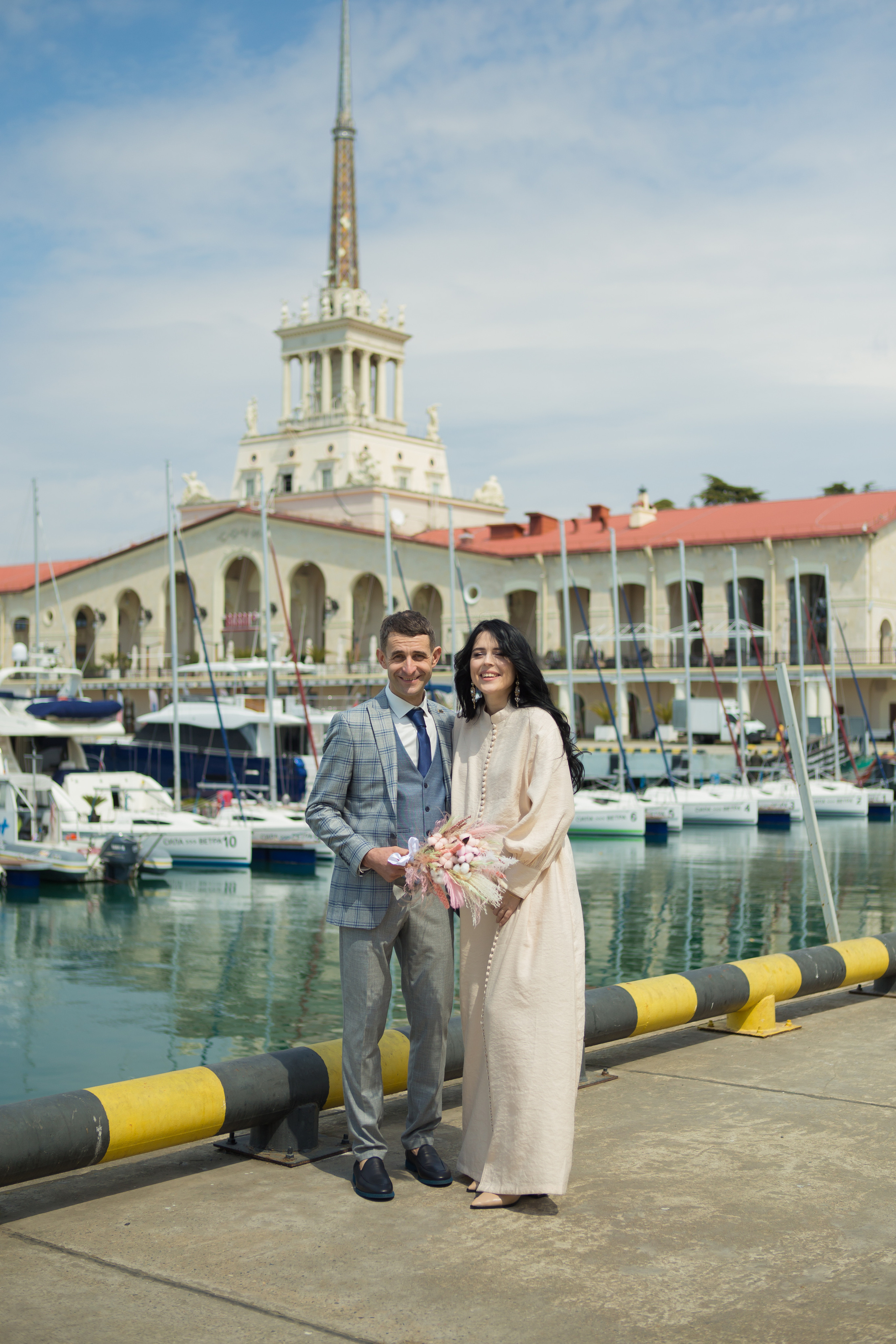 Жених и невеста стоят на фоне здания морского вокзала и моря
