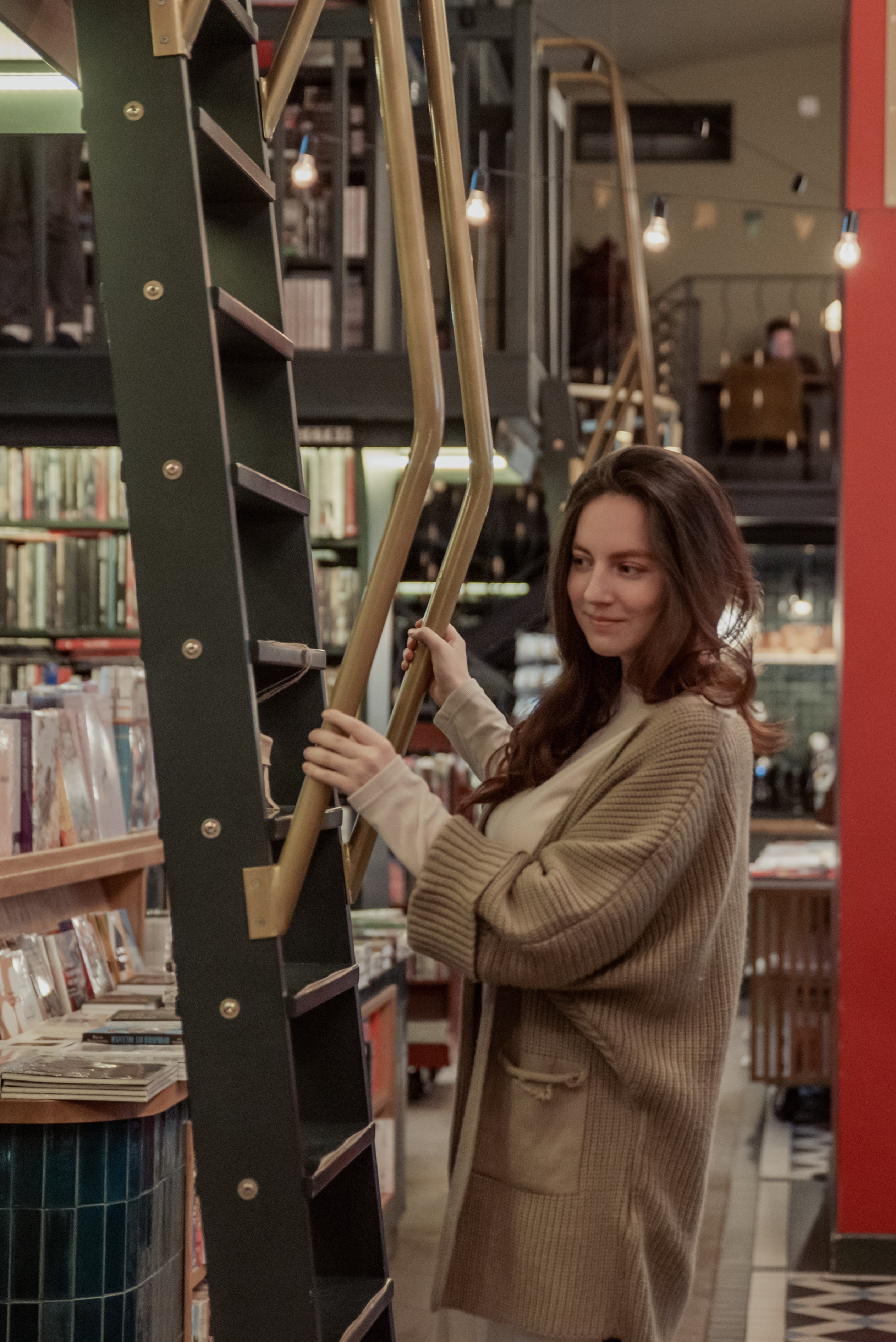 Женщина стоит около высокого книжного шкафа с лестницей и держится за перила.