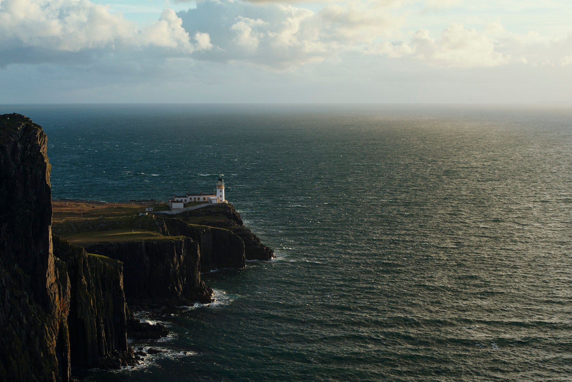 Маяк на мысе Нейст-Пойнт (Neist Point) на краю скалистого утеса острова Скай, Шотландия, с видом на бурные воды моря под частично облачным небом.