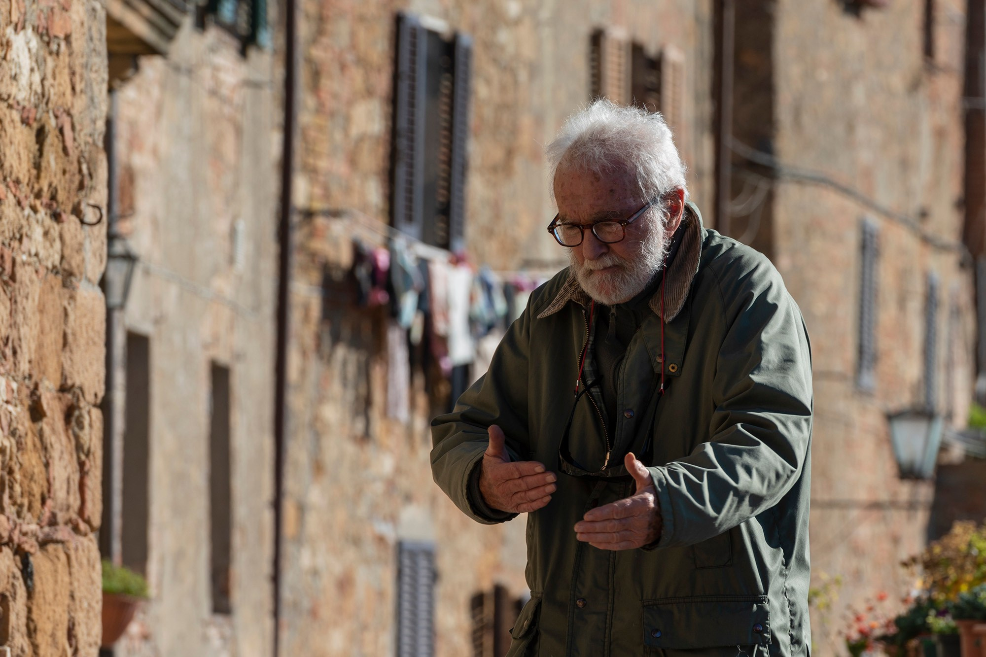 Портрет пожилого мужчины в очках и куртке на солнечной каменной улице в Тоскане, Италия.