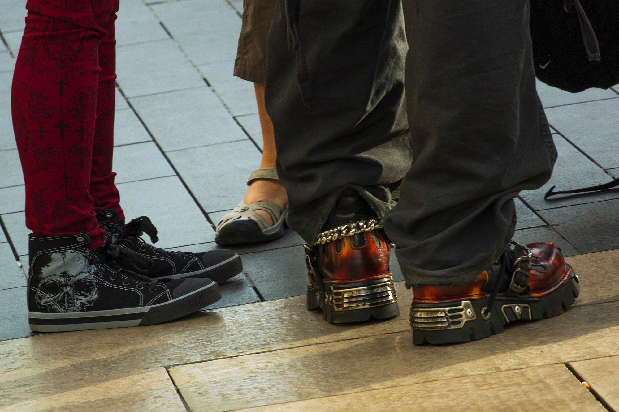 Крупный план ног двух человек в стиле альтернативной моды, показывающий индустриальные готические ботинки с металлическими деталями и кеды с принтом черепа на городской брусчатке.