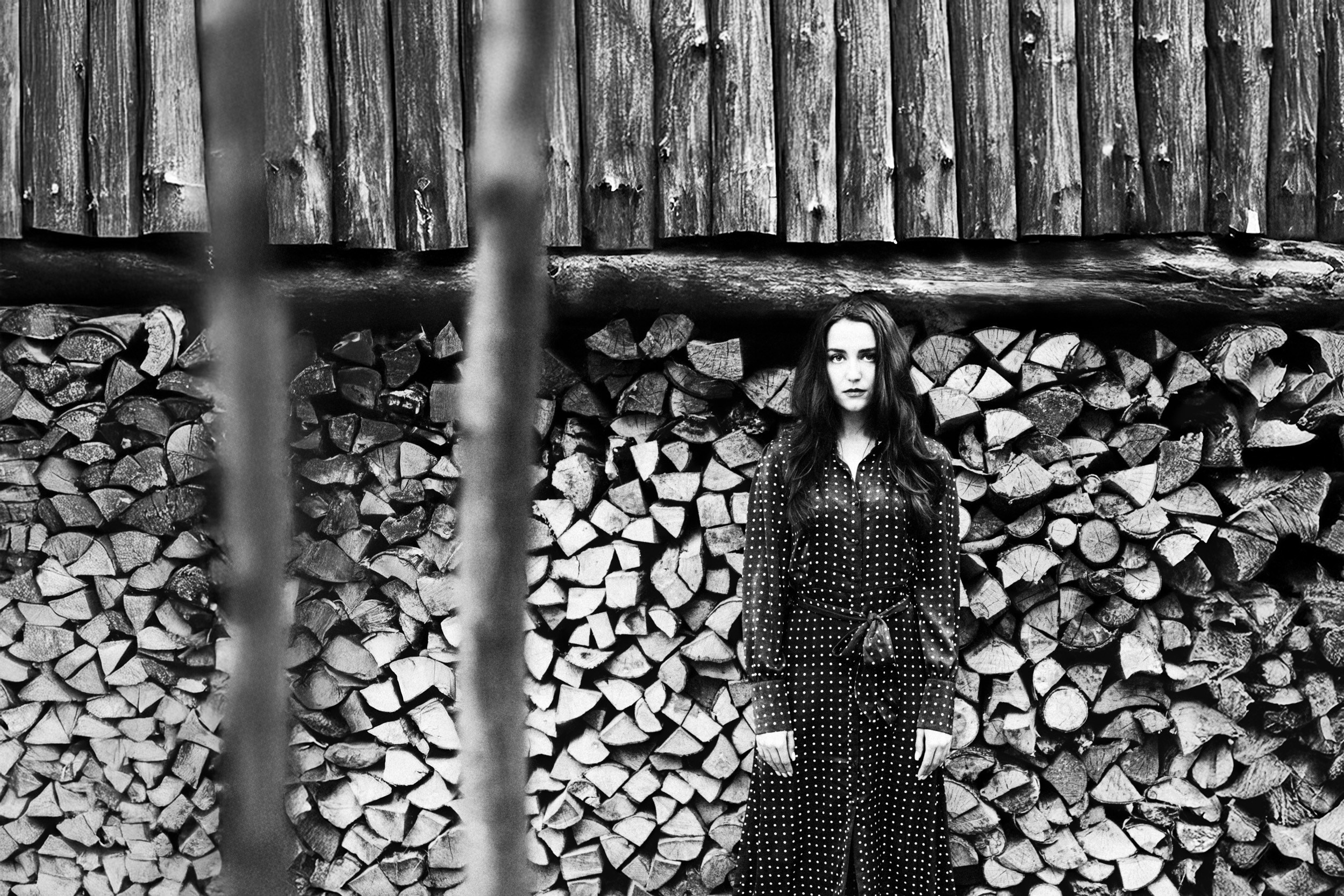 Чёрно-белый портрет женщины в платье в горошек на фоне сложенных дров у деревянной стены