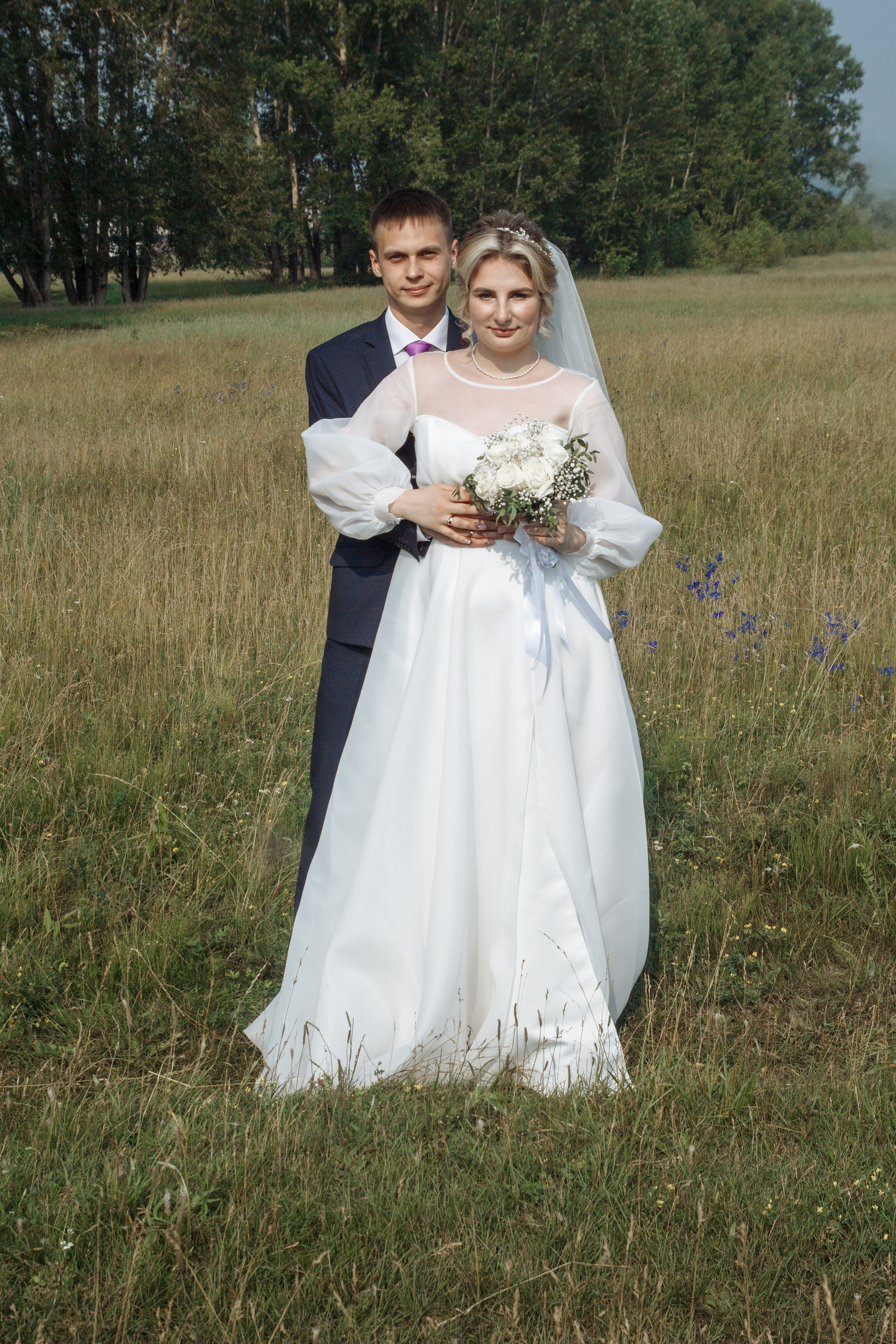 Жених обнимает невесту сзади, оба улыбаются, стоят посреди летнего поля.
