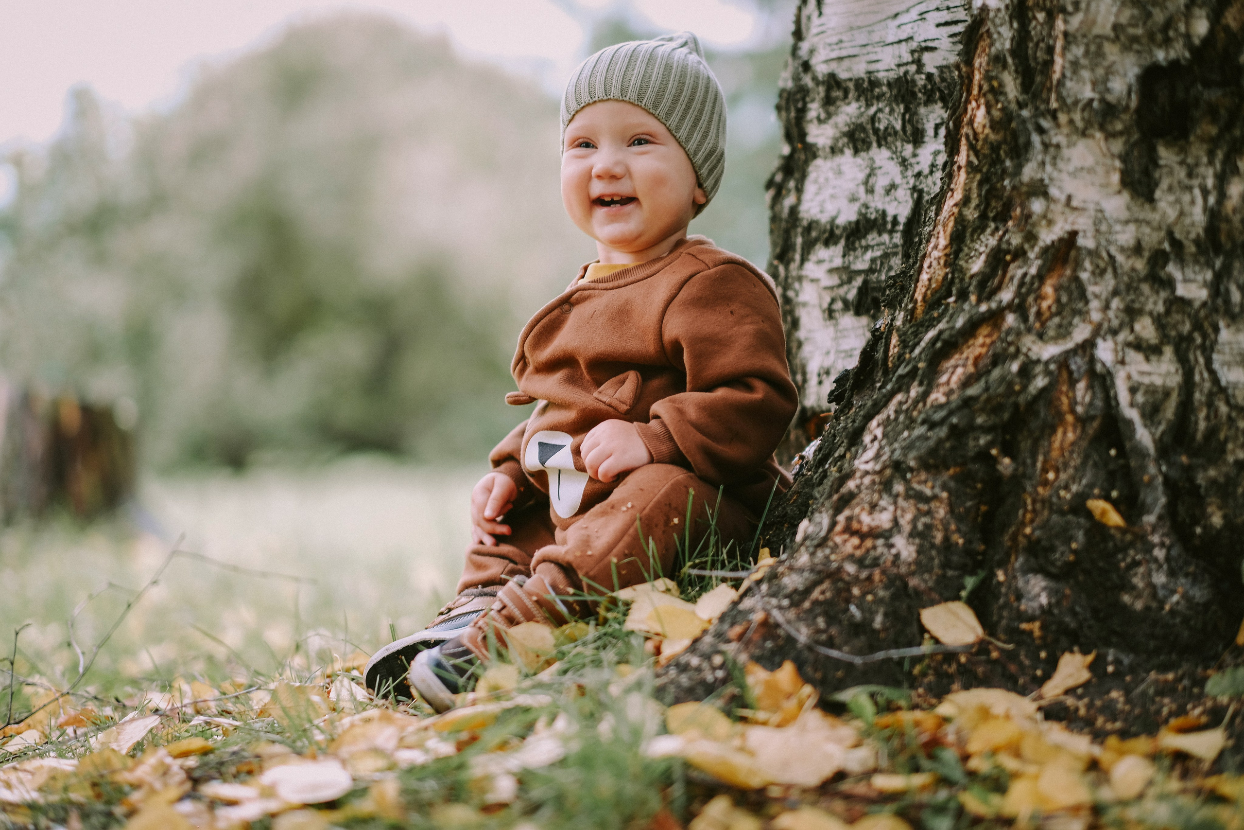 годовалый мальчик малыш у дерева березы осенью в листве и коричневом комбинизоек с шапкой стыковкой сидит и радуется улыбается 