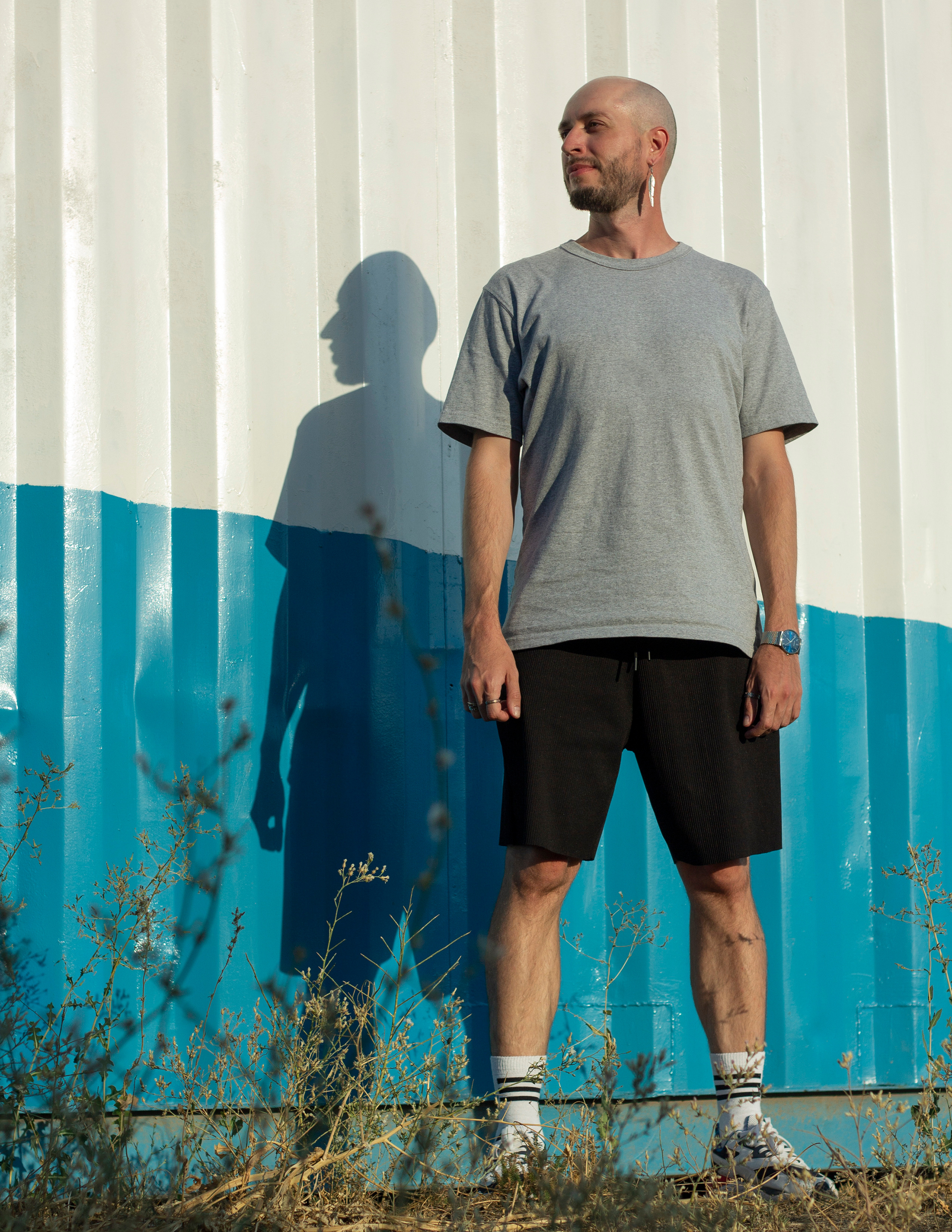 Мужчина в серой футболке и черных шортах стоит на фоне бело-голубой стены и смотрит вправо