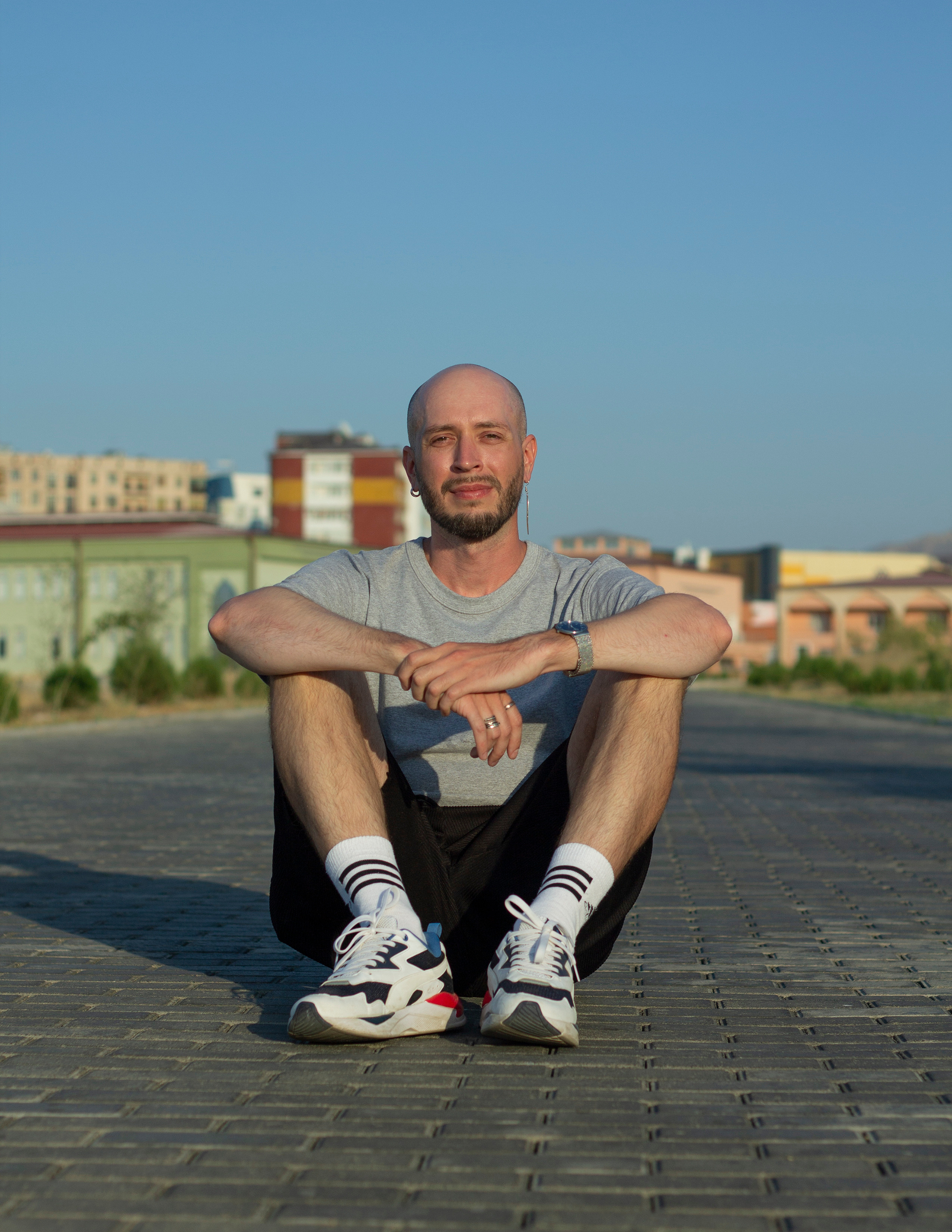 Молодой человек сидит на дорожке на фоне зданий скрестив руки на коленях