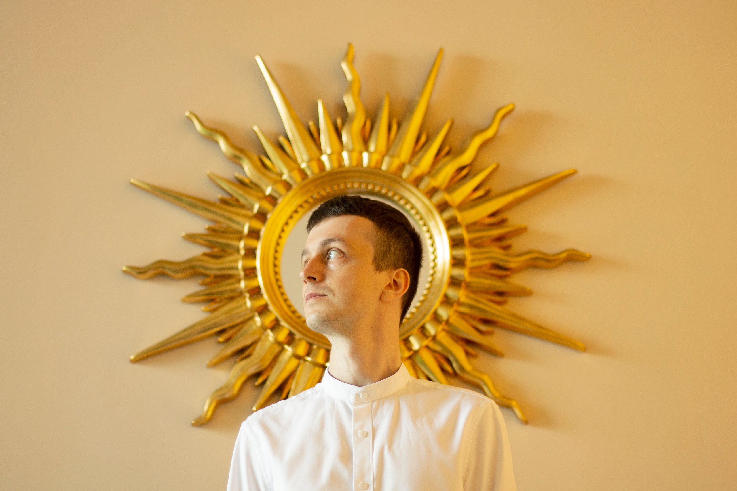 Мужчина в белой рубашке стоит на фоне зеркала в форме солнца 