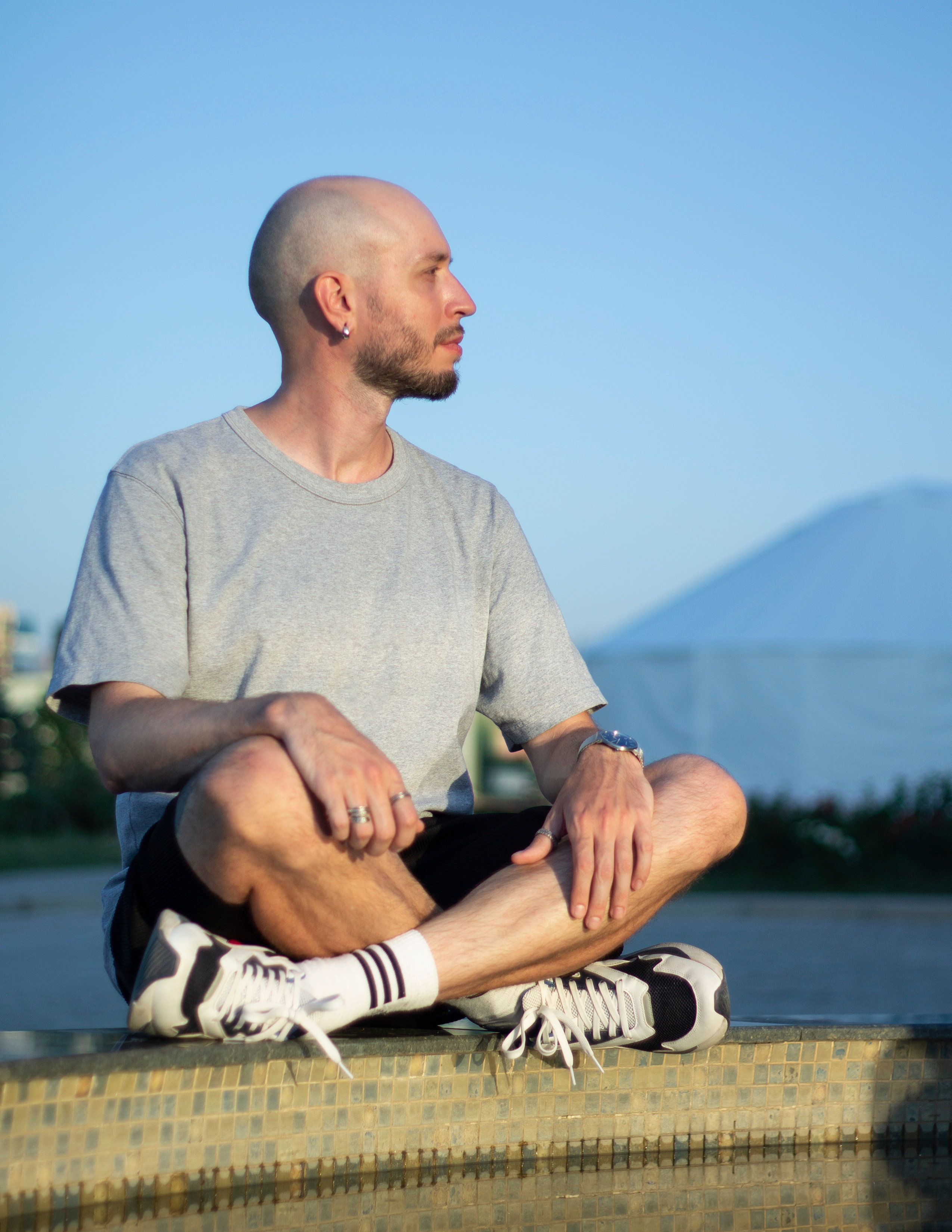 Лысый парень с серьгой в ухе сиди по-турецки на фоне гор и смотрит влево