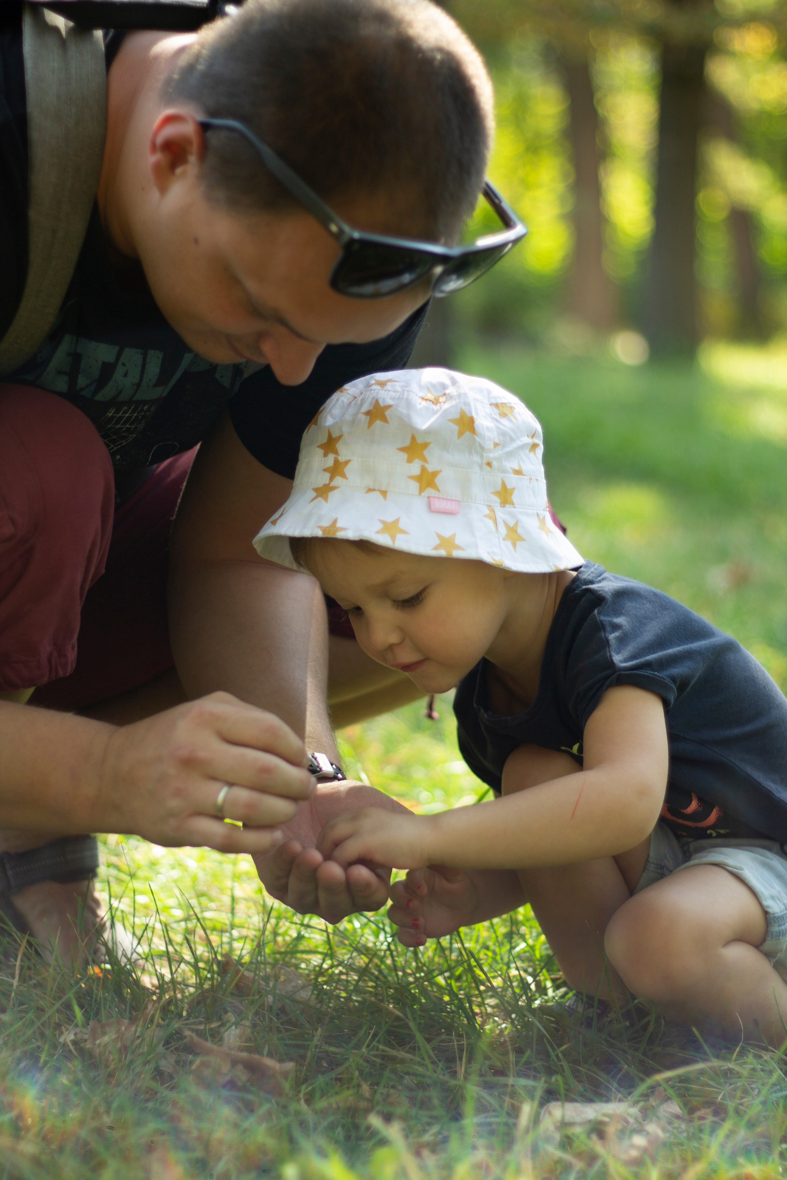 Папа на корточках показывает маленькой дочке в белой панамке кузнечика которого он поймал в ладони