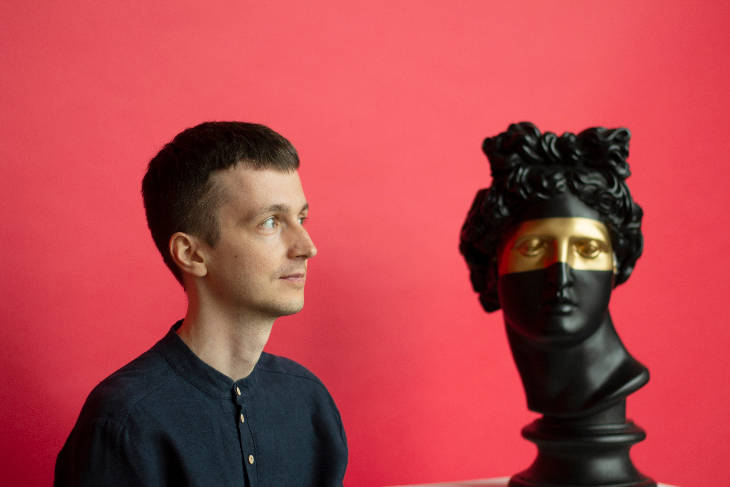 Парень в черной рубашке с черными волосами на красном фоне стоит рядом с черной статуей с золотыми глазами