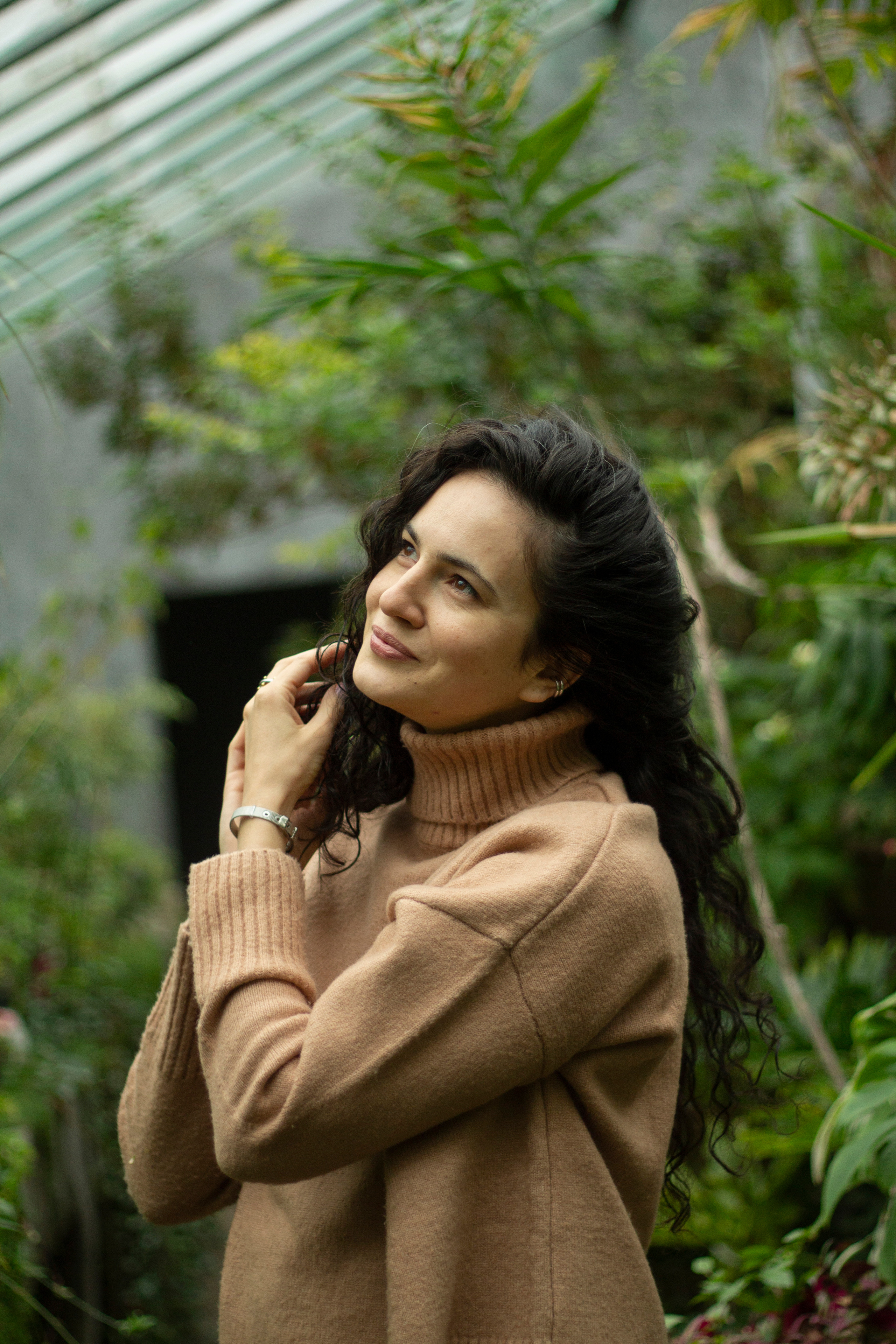 Девушка в свитере стоит в оранжерее и трогает свои распущенные кудрявые темные волосы