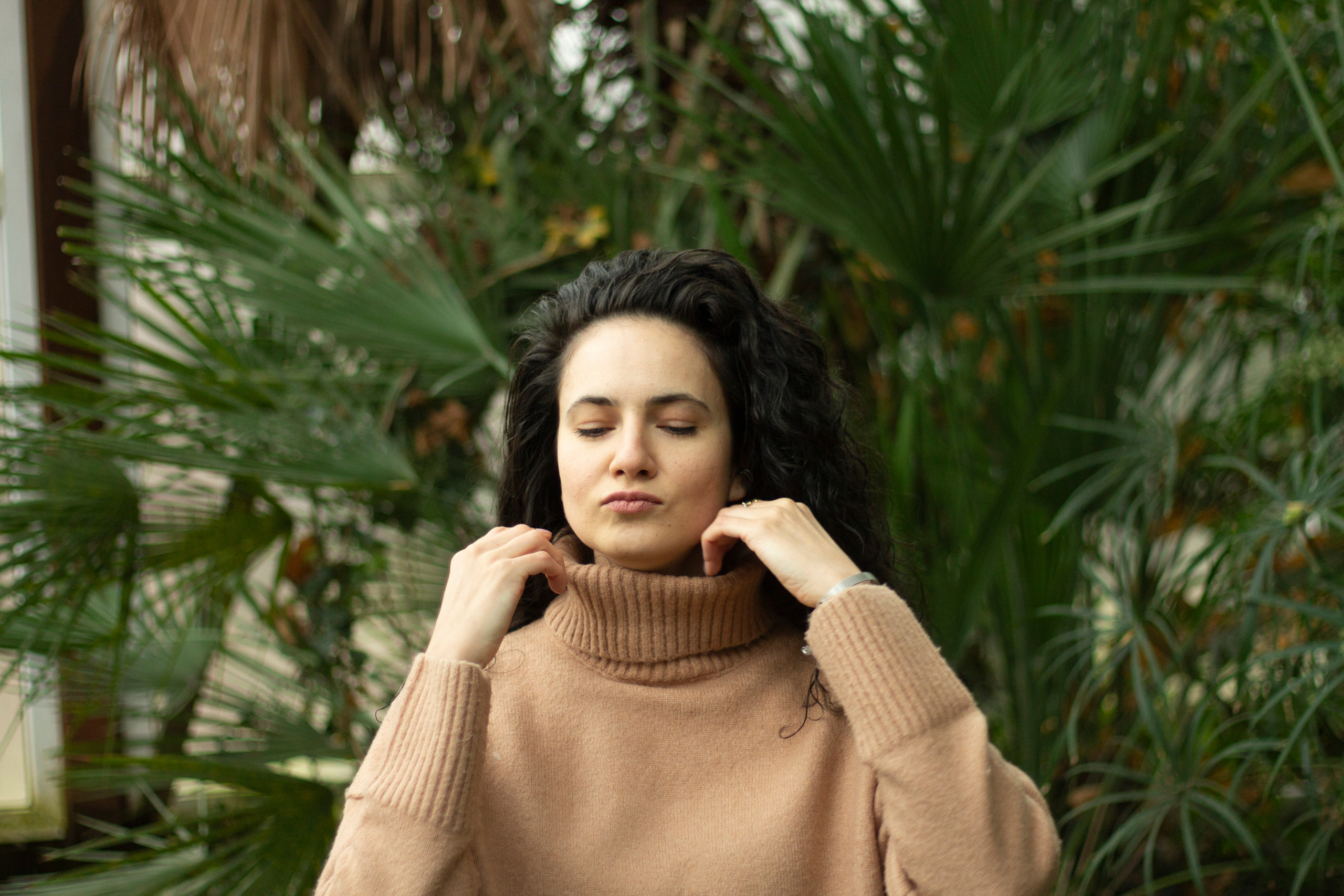 Девушка в оранжерее поправляет воротник бежевого свитера