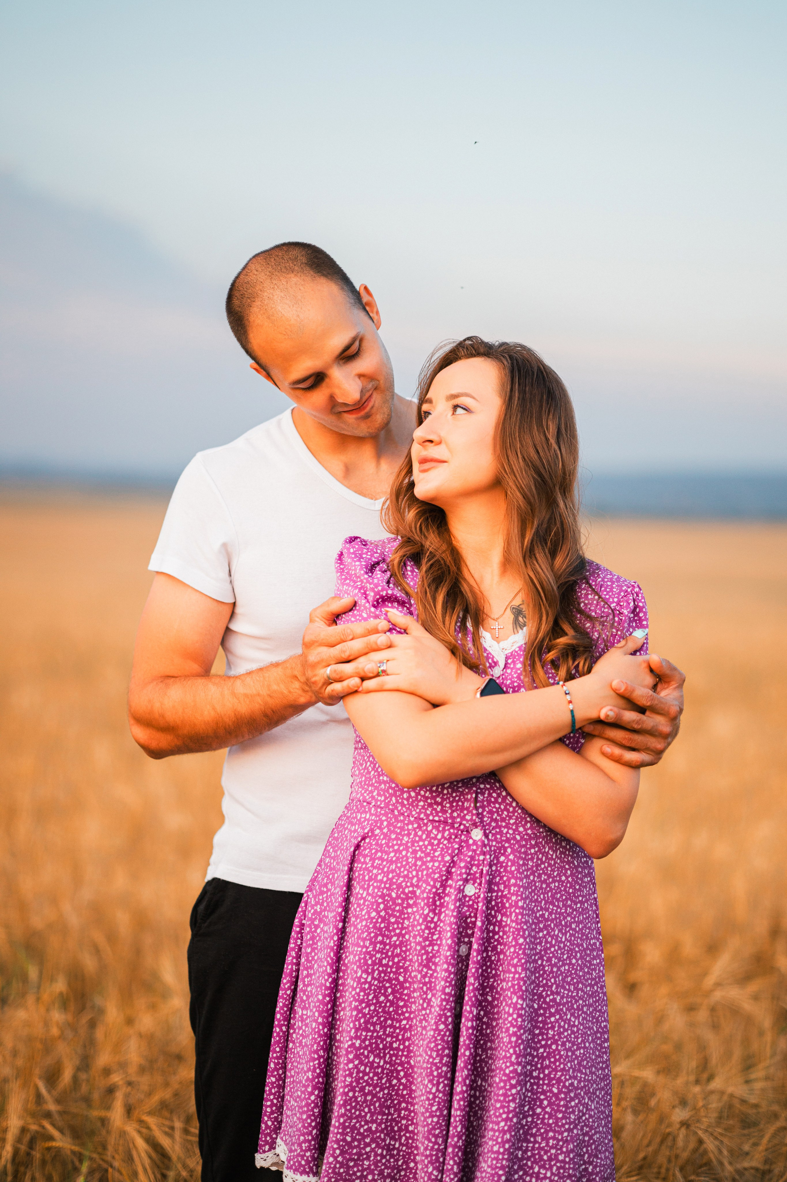 Папа целует маму в щеку, дочка сидит рядом на пледе в поле при закате.
