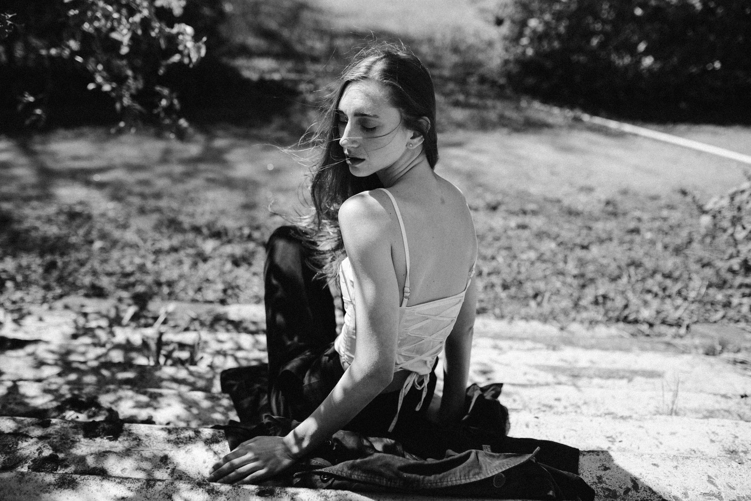 Девушка в белом корсете с обнаженными плечами сидит на каменных ступенях под солнцем