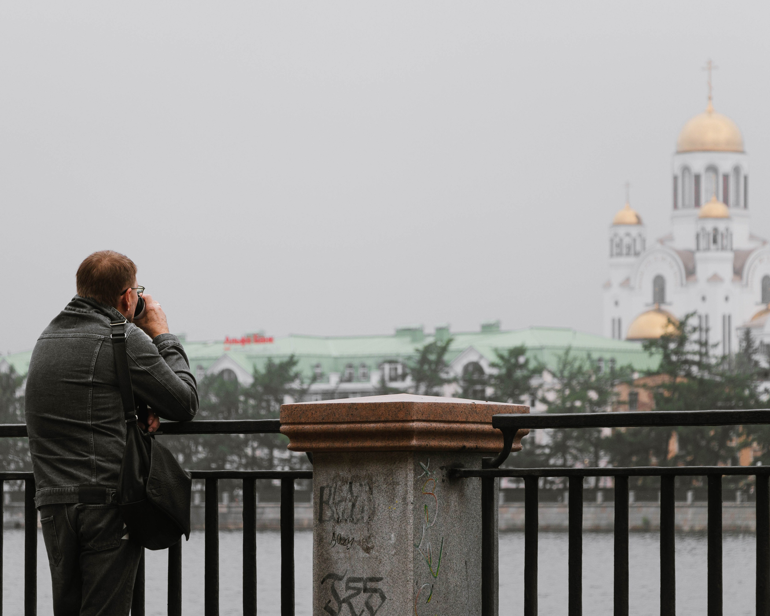 Мужчина с чашкой кофе стоит у забора и смотрит на Храм на крови