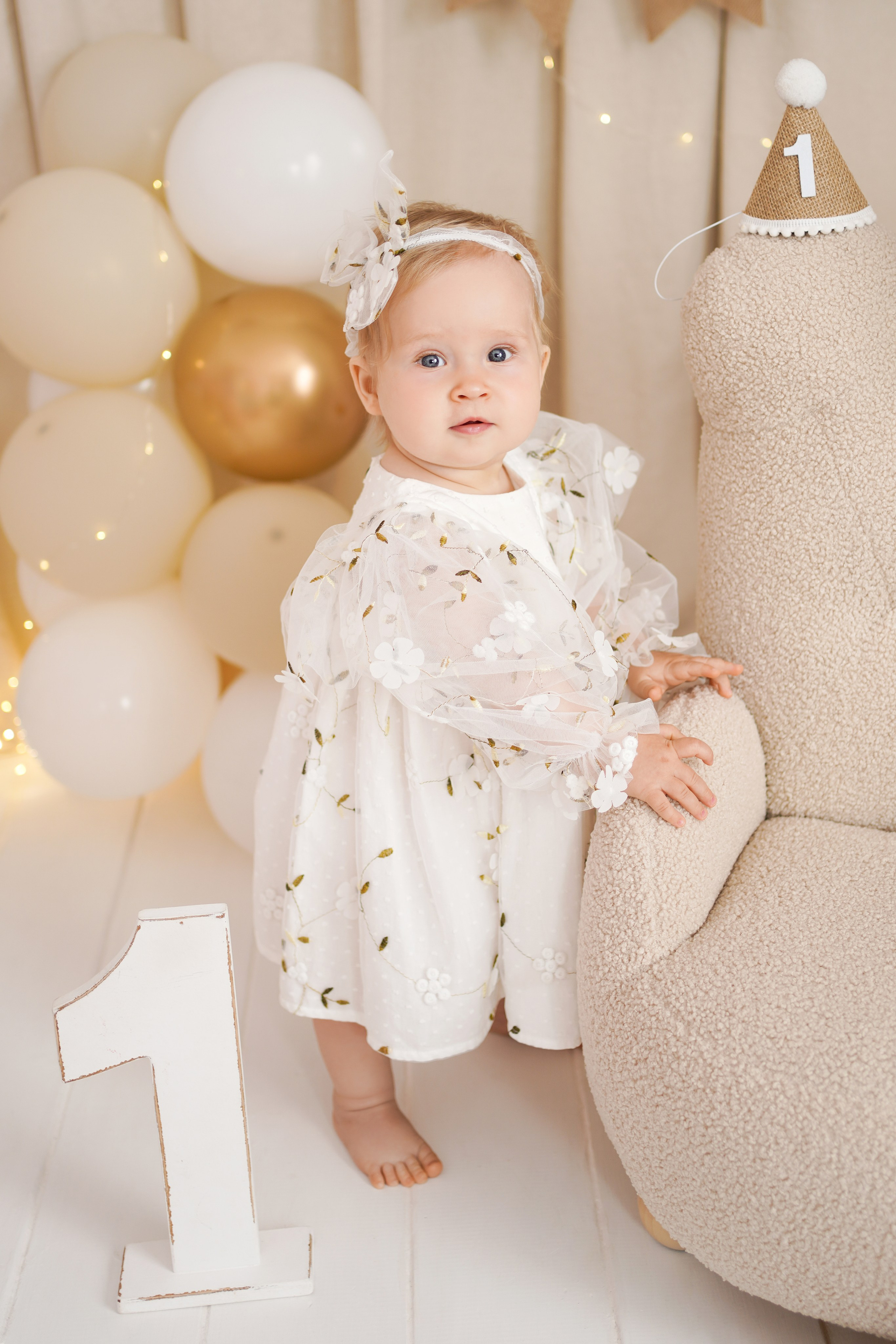 Малышка отмечает Первый годик, она в нарядной одежде, рядом стоит единичка, много воздушных шаров.