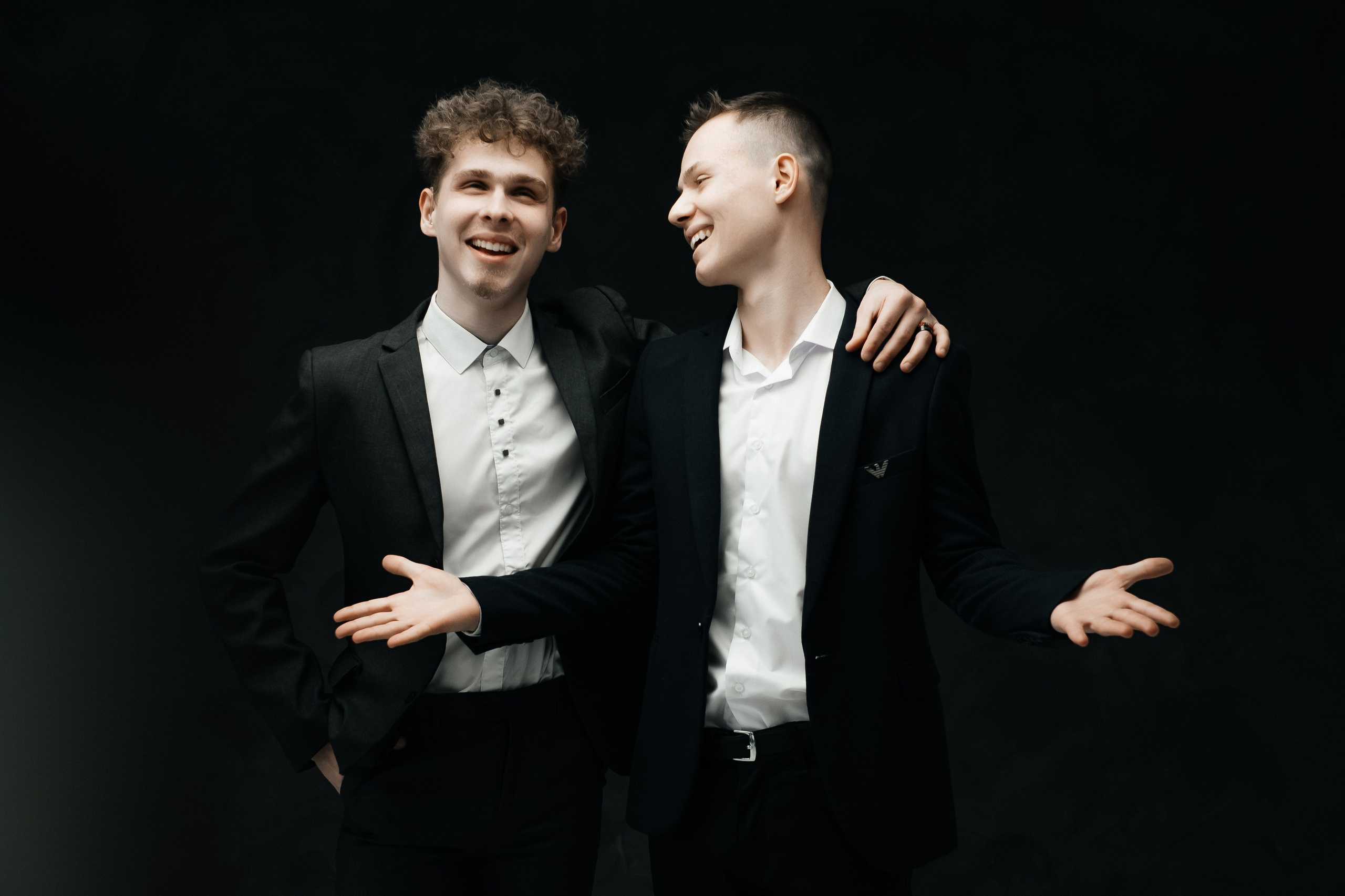 Два молодых человека два парня в черных костюмах и белых рубашках обнимаются друзья и улыбаются 