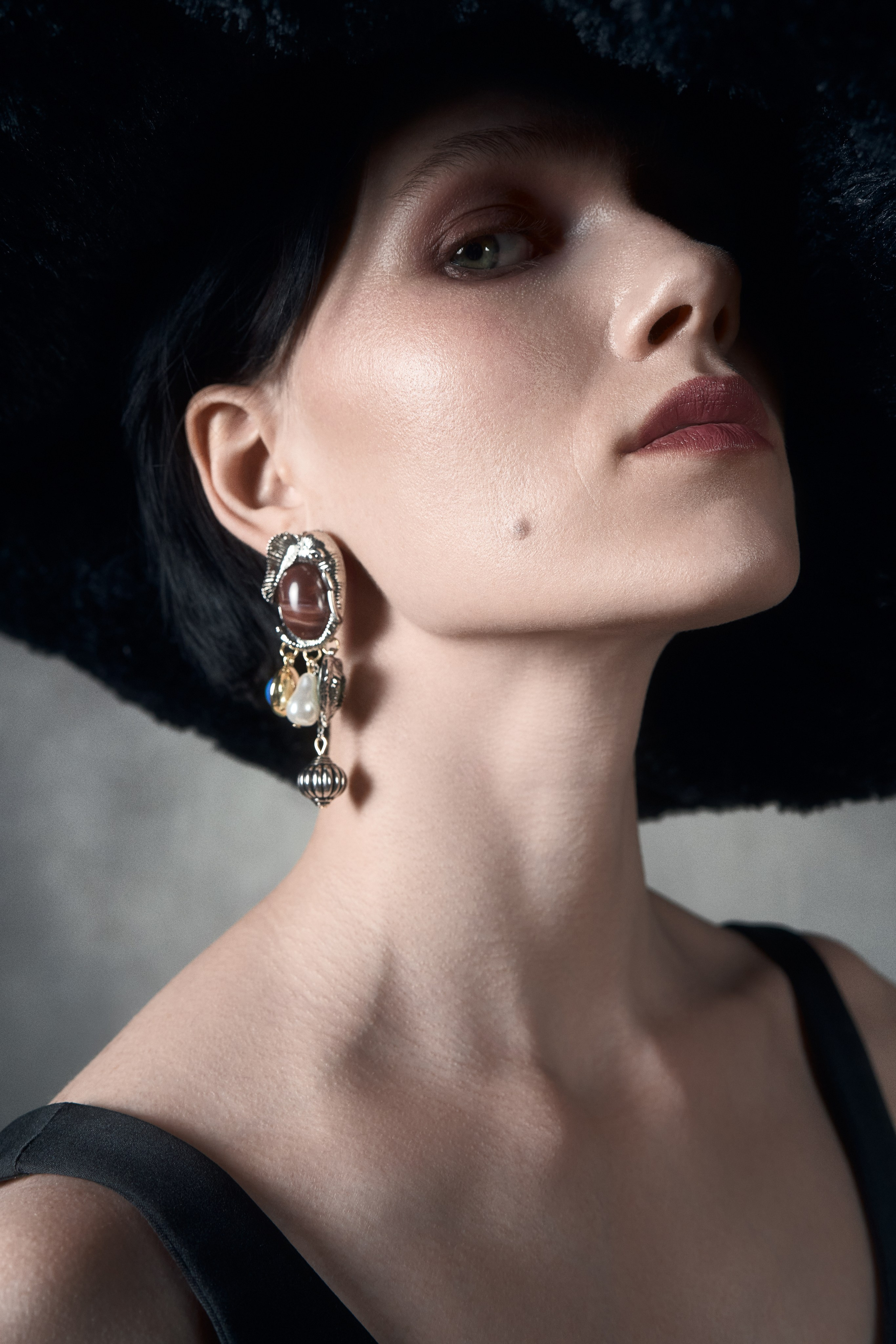 Крупный портрет модели в широкой чёрной шляпе, художественная fashion-фотография с акцентом на украшения и характер образа
