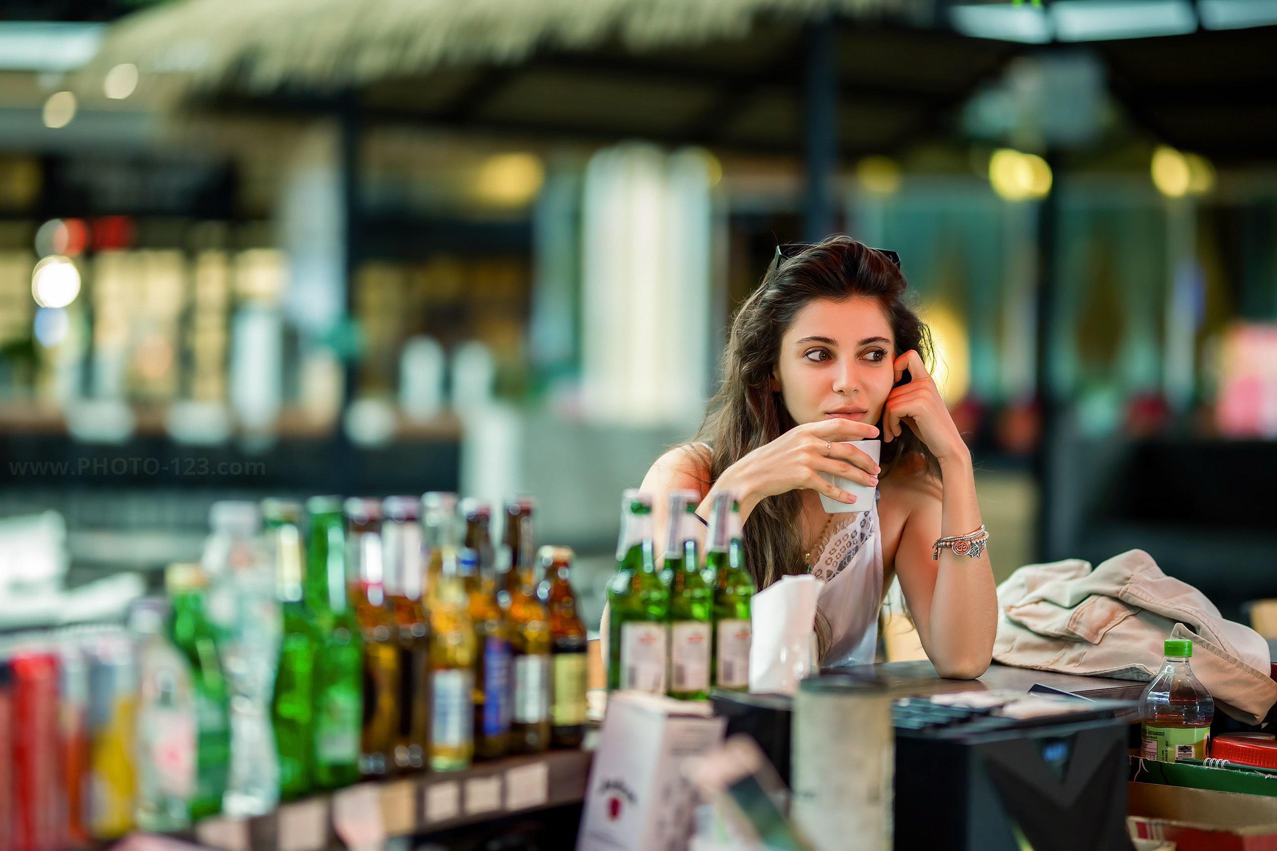 Lifestyle portrait of a woman sitting at a tropical bar counter, holding a white cup and looking sideways; surrounded by colorful bottles of beer and liquor, with thatched roof and soft background blur; editorial-style image captured in Phu Quoc, highlighting relaxed mood and vibrant textures