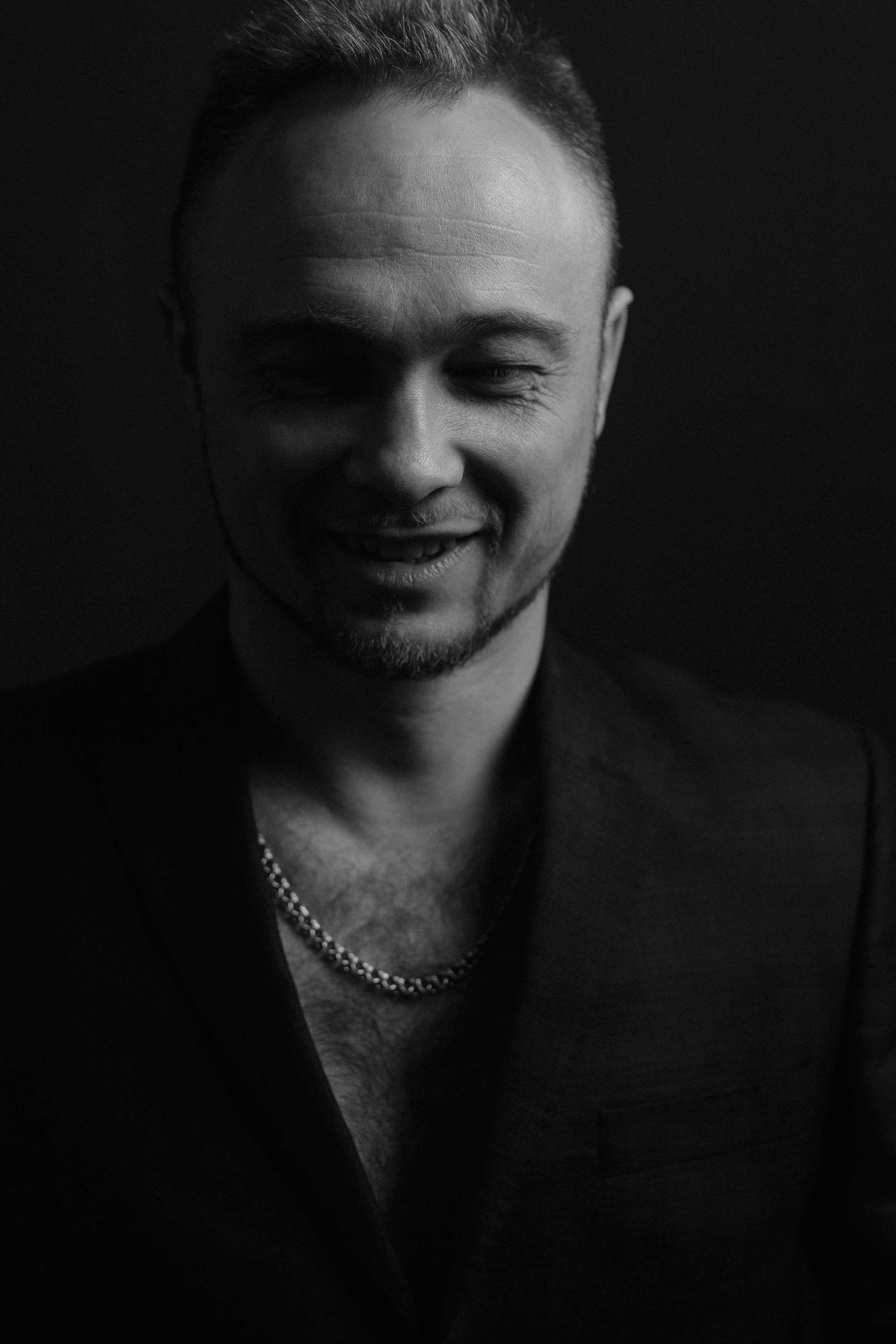 Мужчина в тёмном пиджаке с цепочкой на шее слегка улыбается, глядя вниз. Чёрно-белый стиль добавляет снимку глубины и контраста