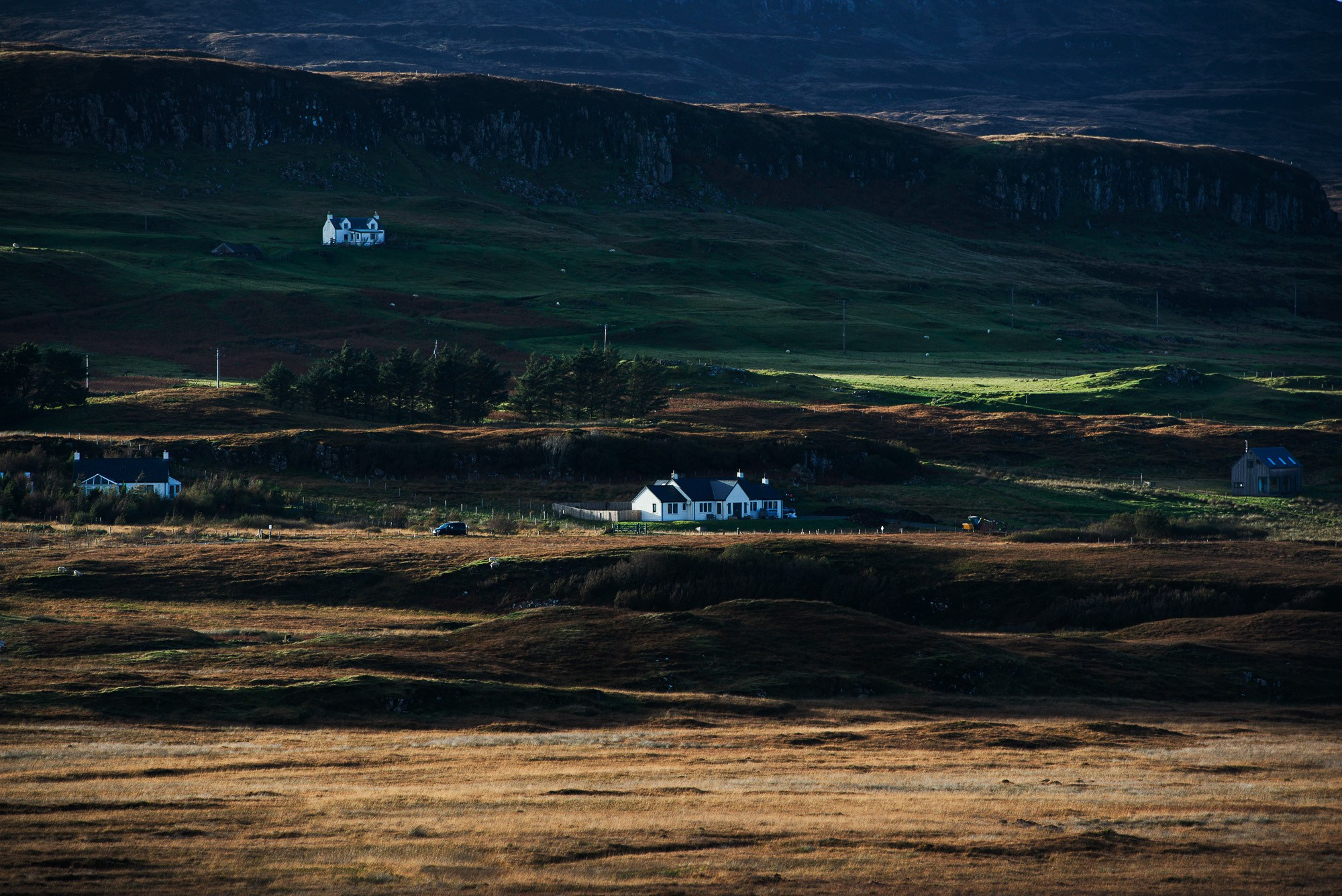 Деревенские дома в долине на острове Скай, Шотландия, с сухими желтыми полями на переднем плане и темными зелеными холмами на заднем плане под драматичным солнечным светом.