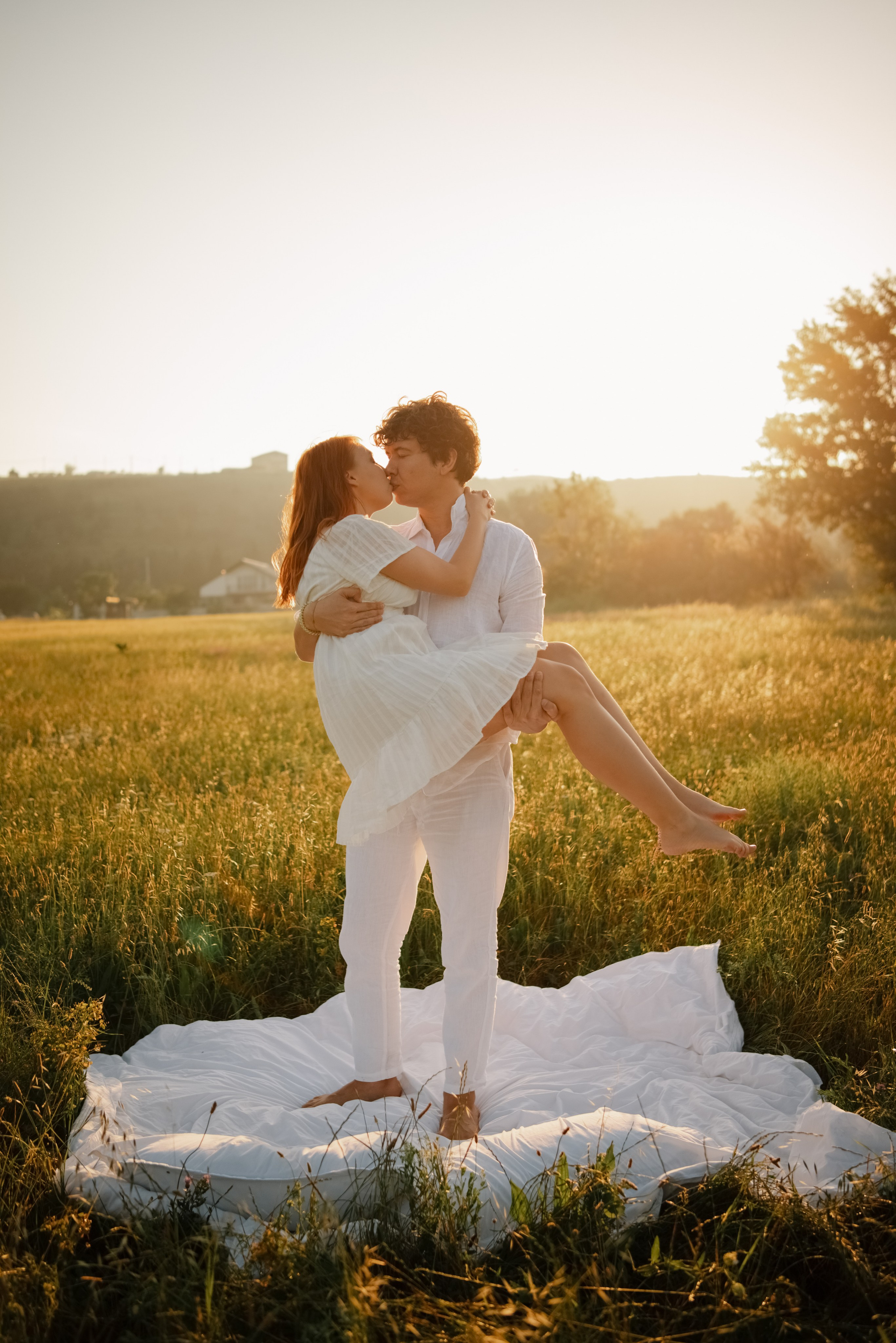 Влюбленные в белой одежде в поле на фоне рассвета. Мужчина держит девушку на руках и целует