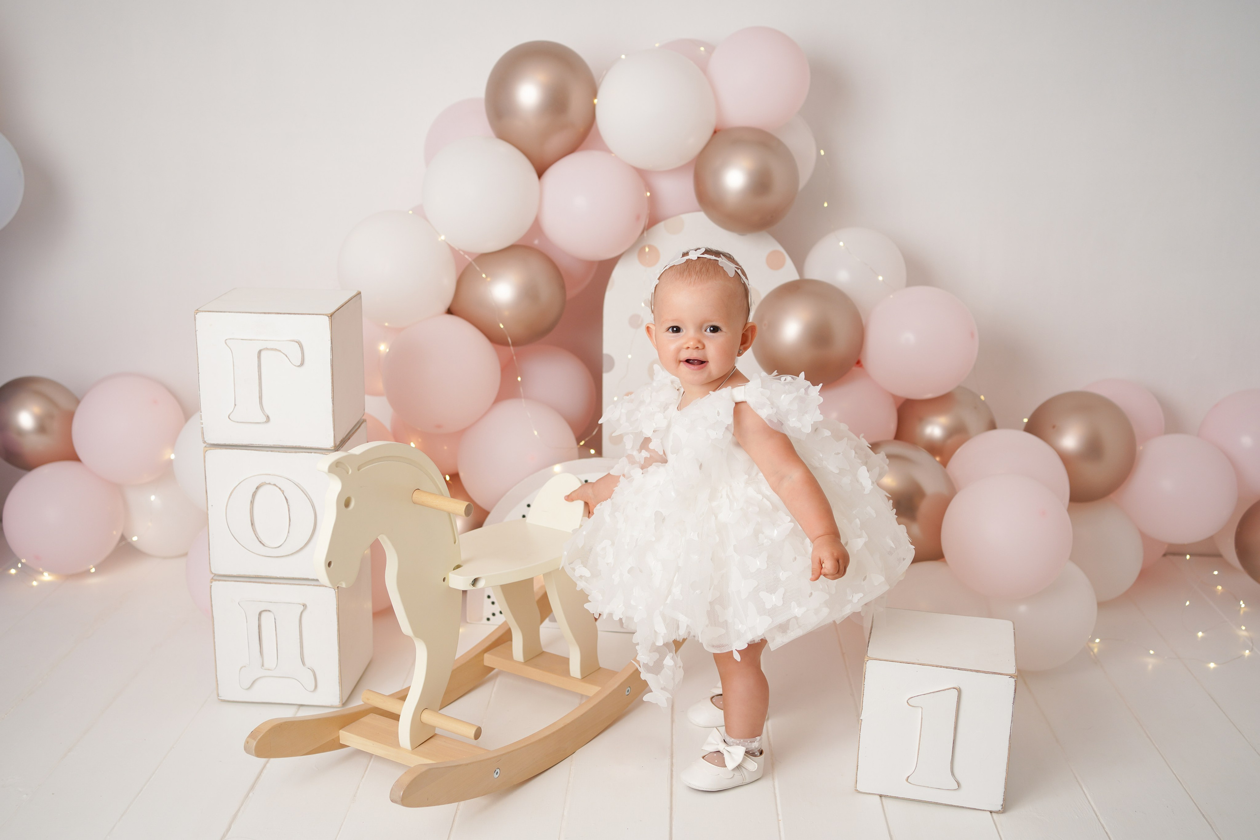 На фото малышка скачет на лошадке. рядом много розовых шаров и  кубики год.