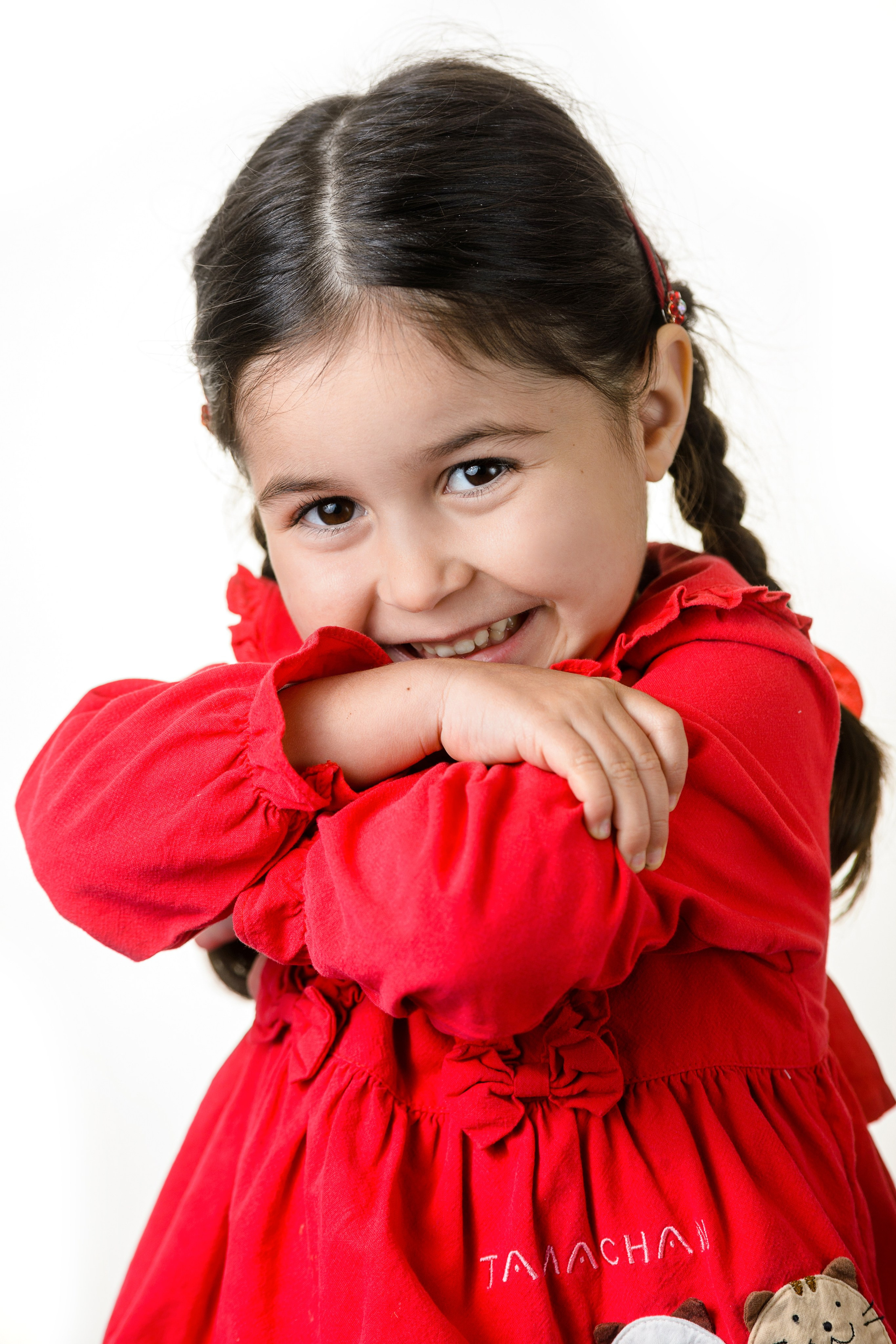 Девочка в красном платье закрывает лицо руками и улыбается в студийном портрете на белом фоне.