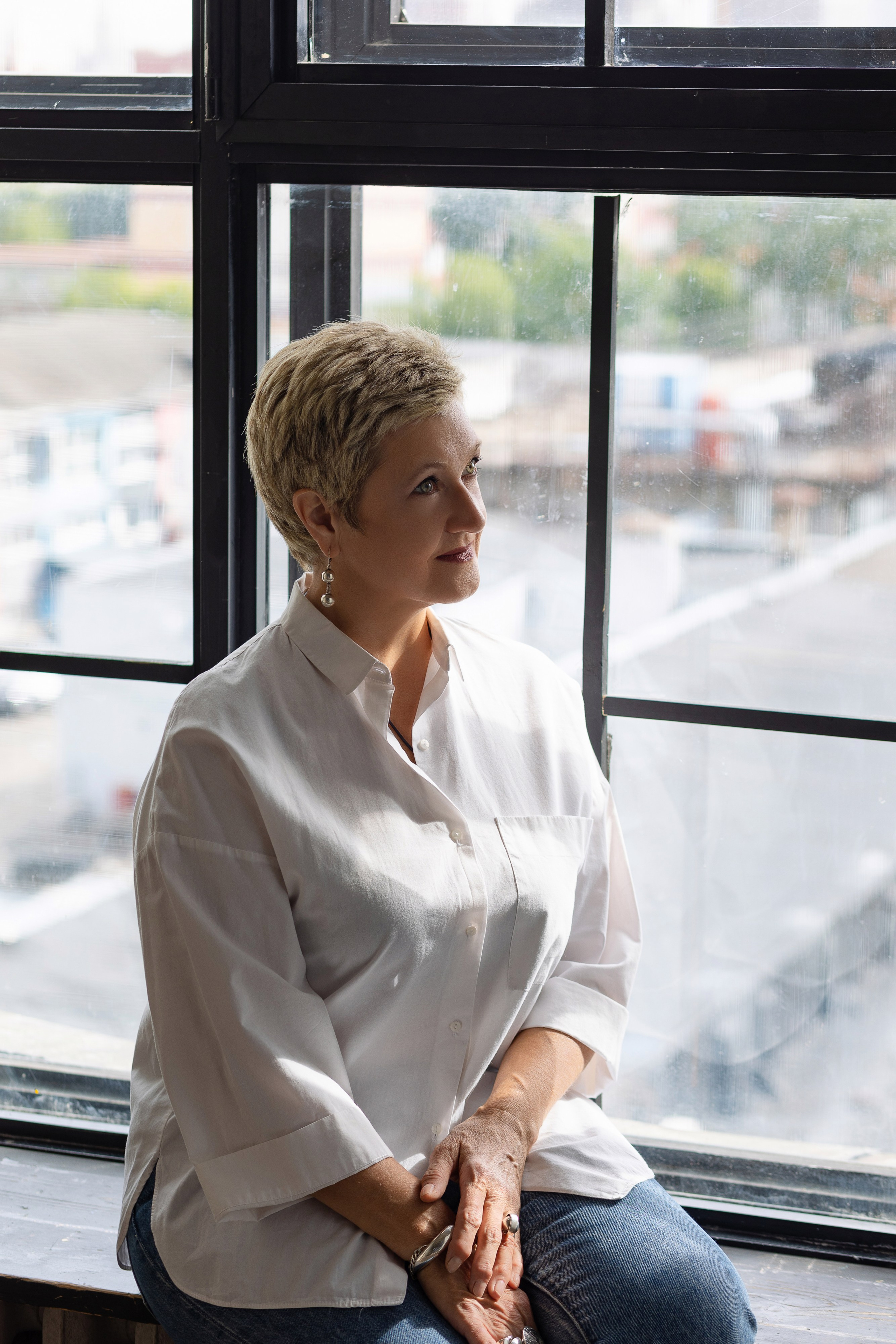 Женщина сидит на подоконнике, за окном солнечный городской пейзаж.