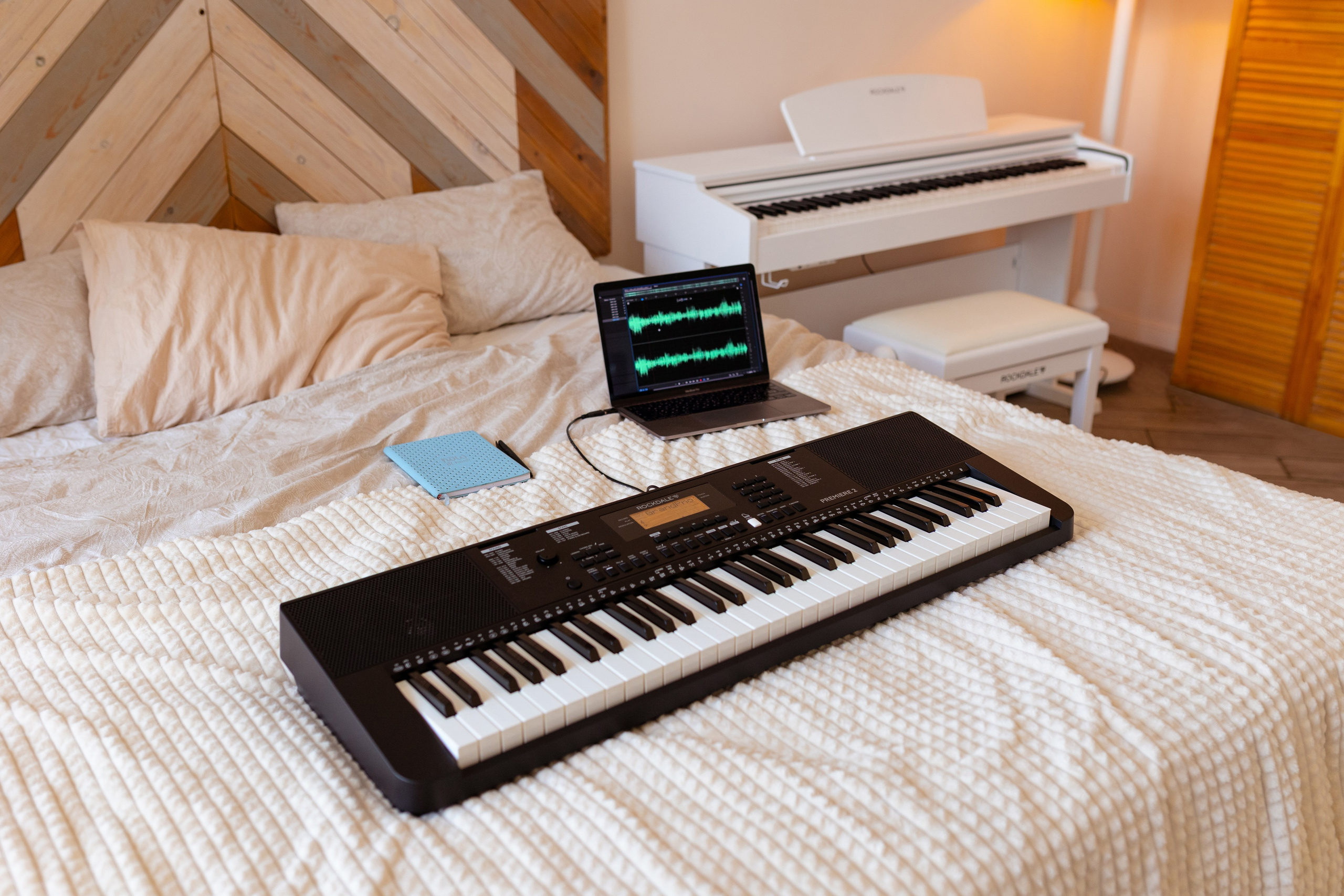 На белой кровати лежит пианино и ноутбук.