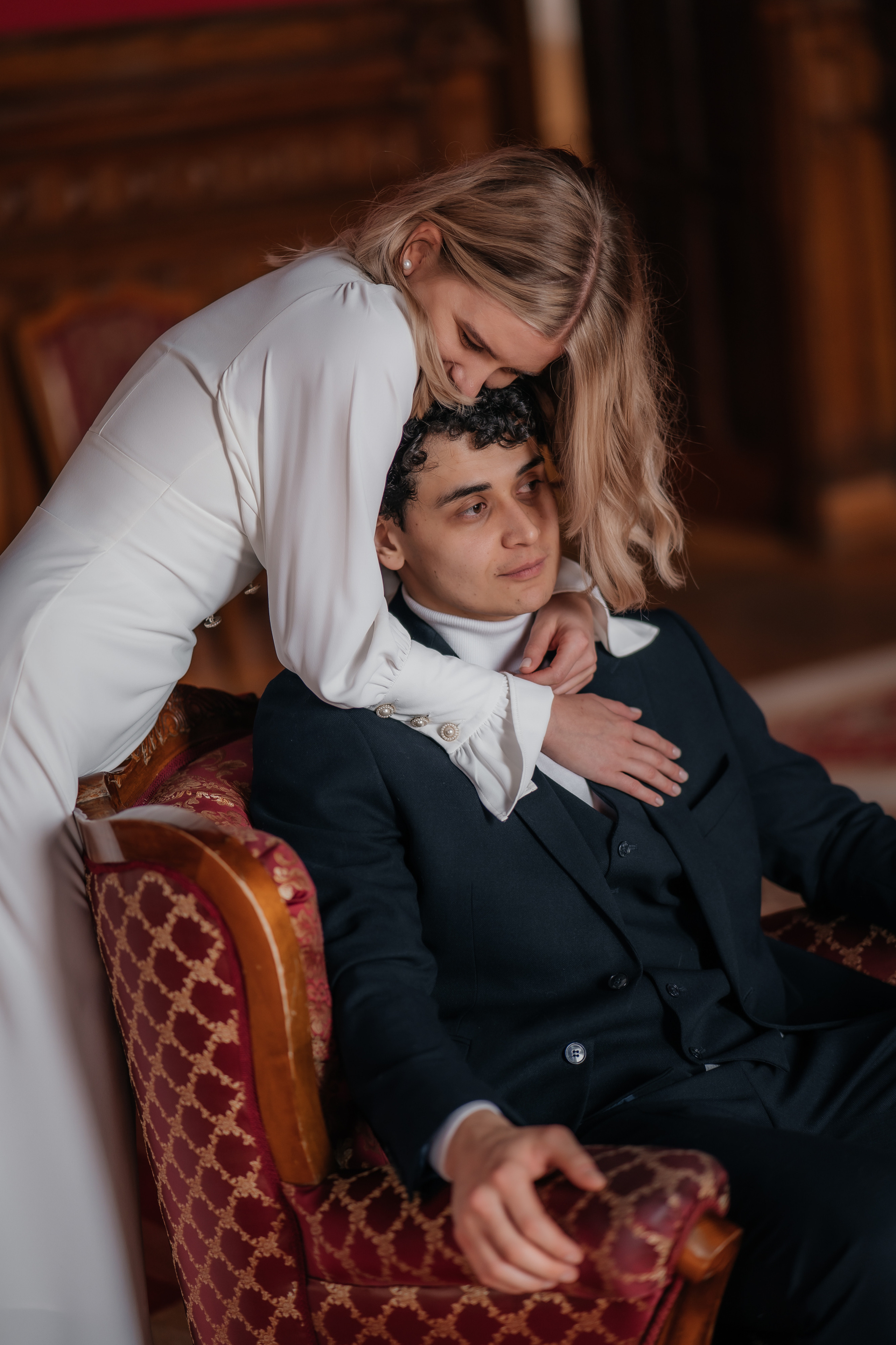 Жених сидит в кресле в красном зале дворца бракосочетания №2, а невеста обнимает его